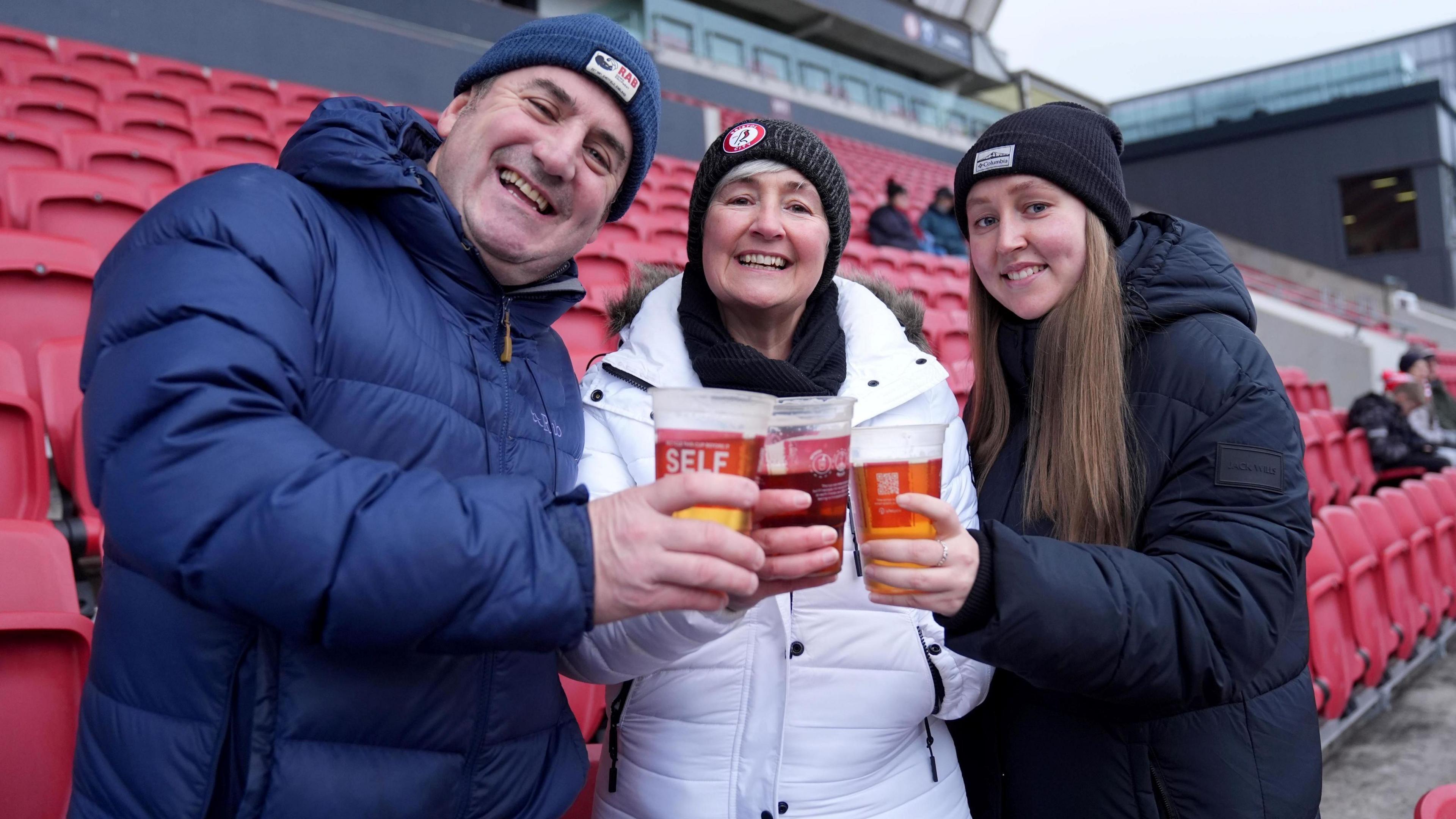 Three fans smile at the camera as they touch their pint glasses together in the stands at Ashton Gate before a game involving Bristol City Women's FC. Two of the group are women and one is a man and they are all dressed in winter clothing