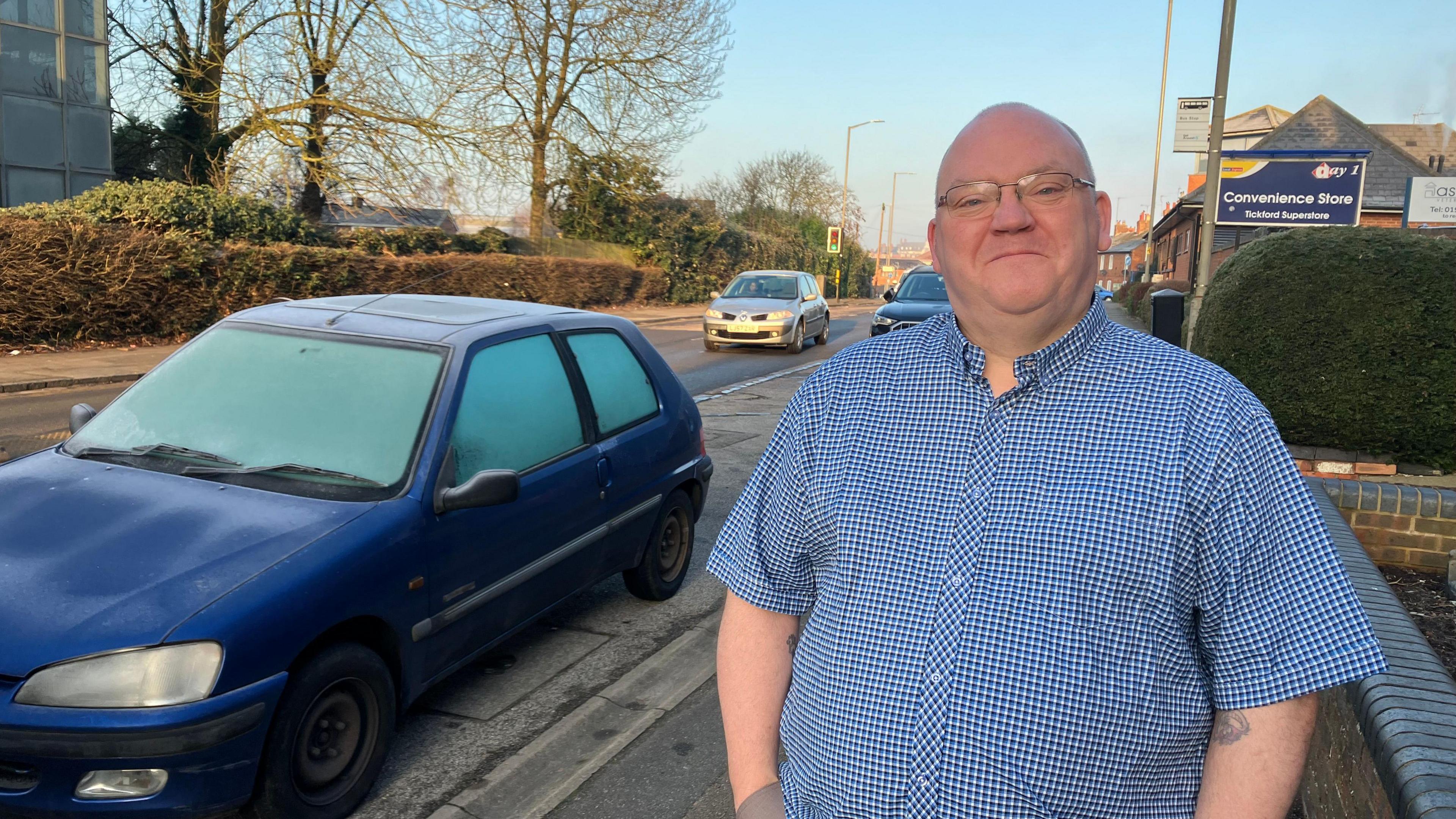 A picture of a man, standing on a pavement next to a road and a blue car. He has his hands in his pocket and is wearing a blue short sleeved shirt. He has glasses on.