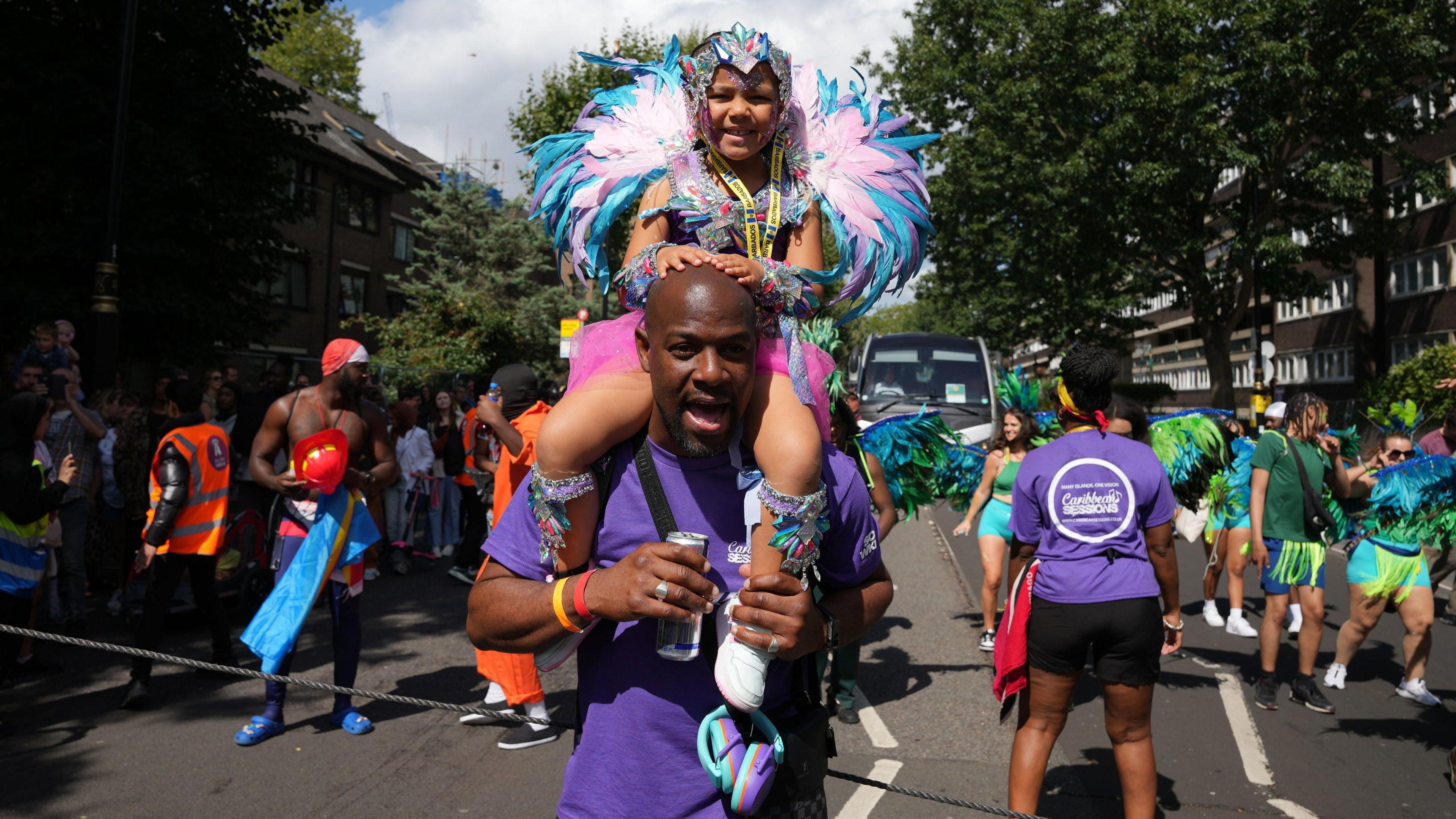 A girl in a feathered costume is carried through the street on her dad's shoulders