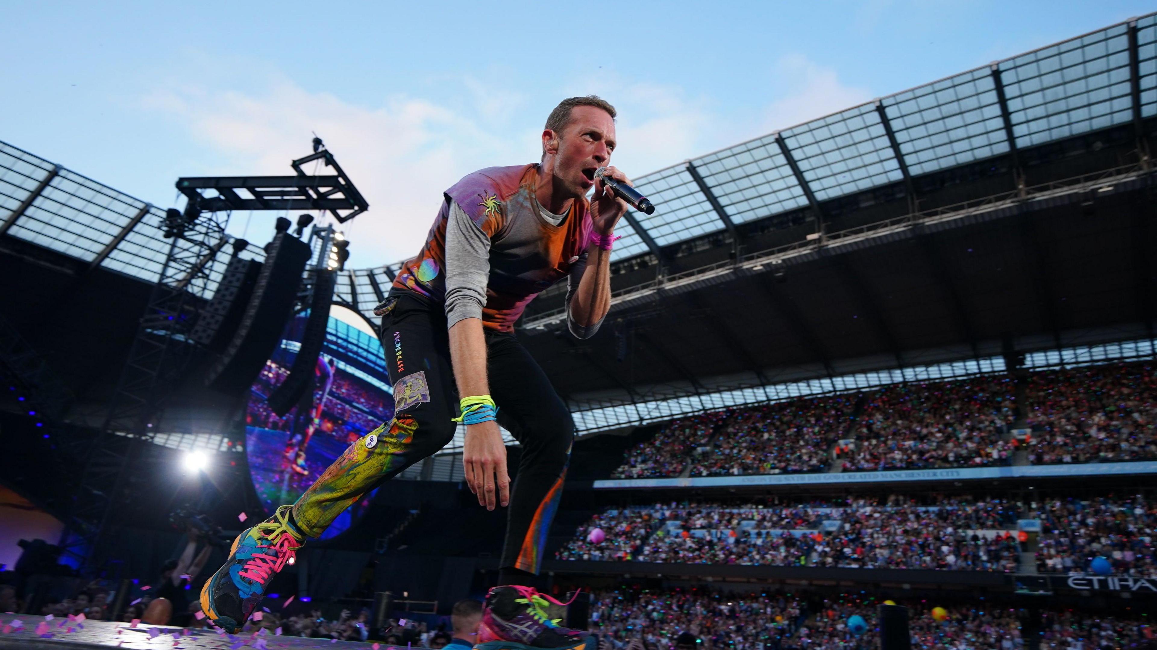 Chris Martin, wearing paint coloured clothes, performs on stage at the Etihad Stadium in Manchester.