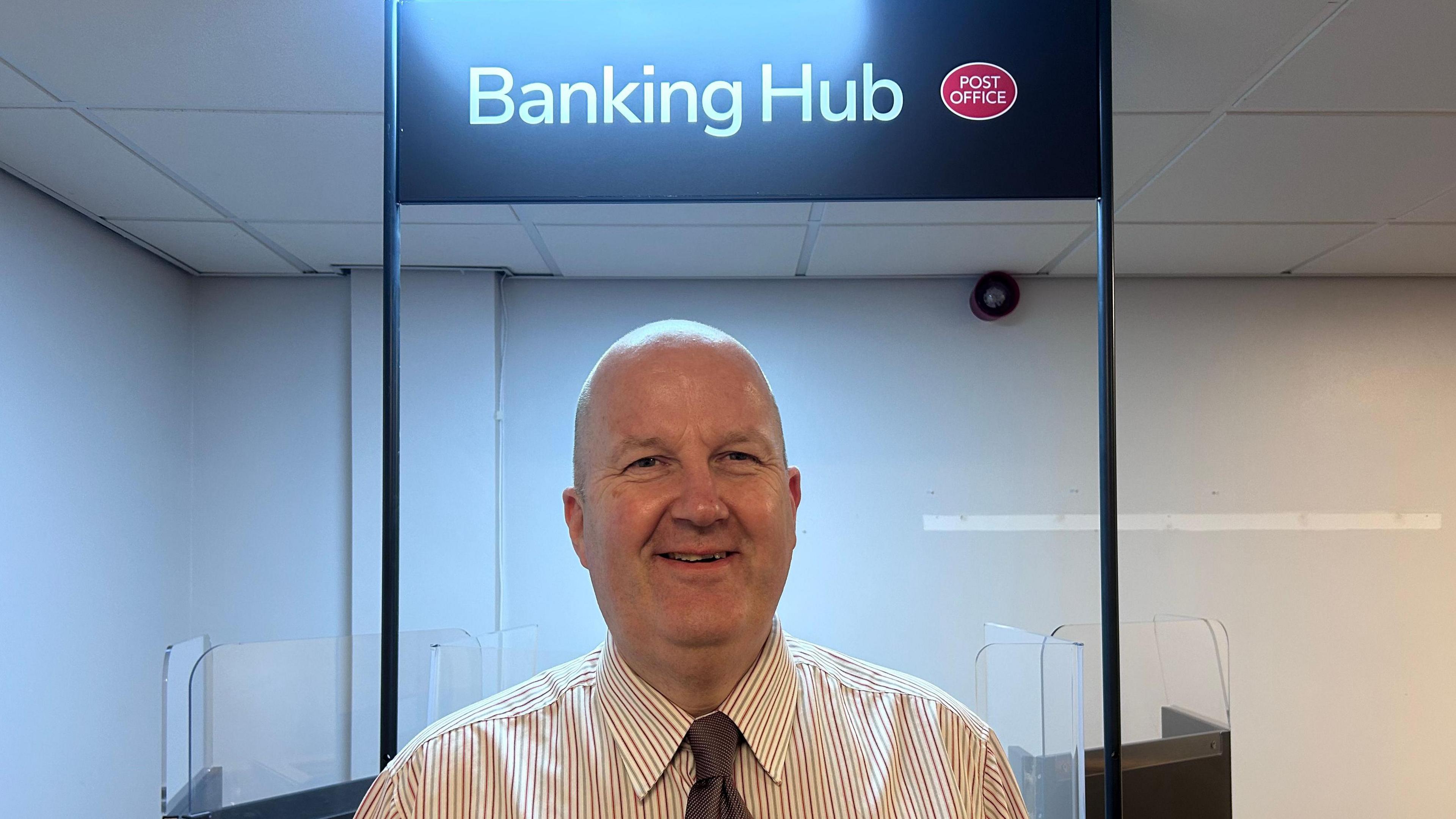 A man in a stripy shirt wearing a tie stood in front of a counter with a sign above his head reading: Banking Hub.