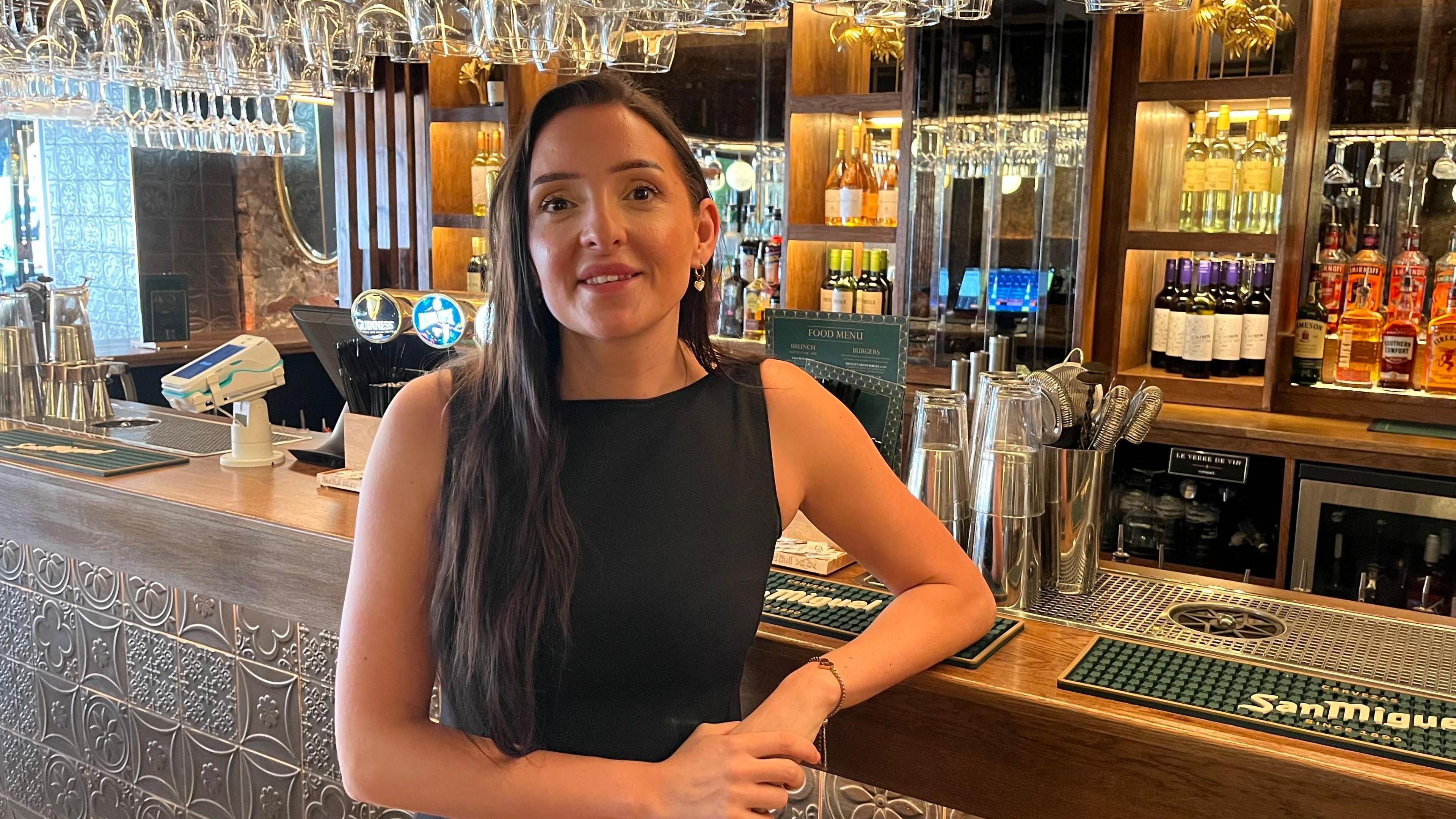 A woman with long dark hair and wearing a black dress stands in front of a bar. Bottles of alcohol are displayed on shelves in the background. Rows of wine glasses are also hanging from the ceiling.