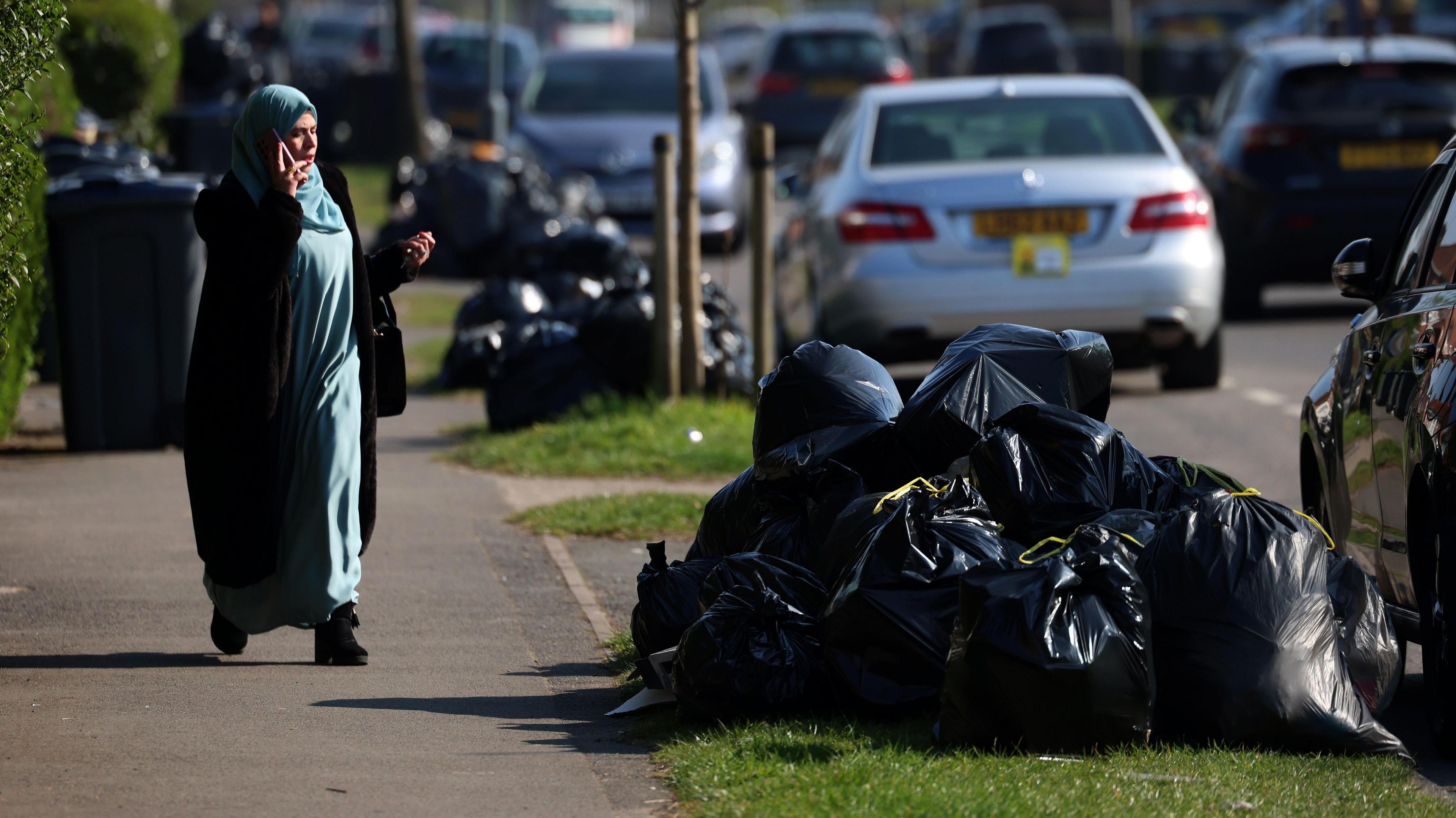 A woman walks down the street, past piles of black bin bags and parked cars, while talking into a mobile phone.