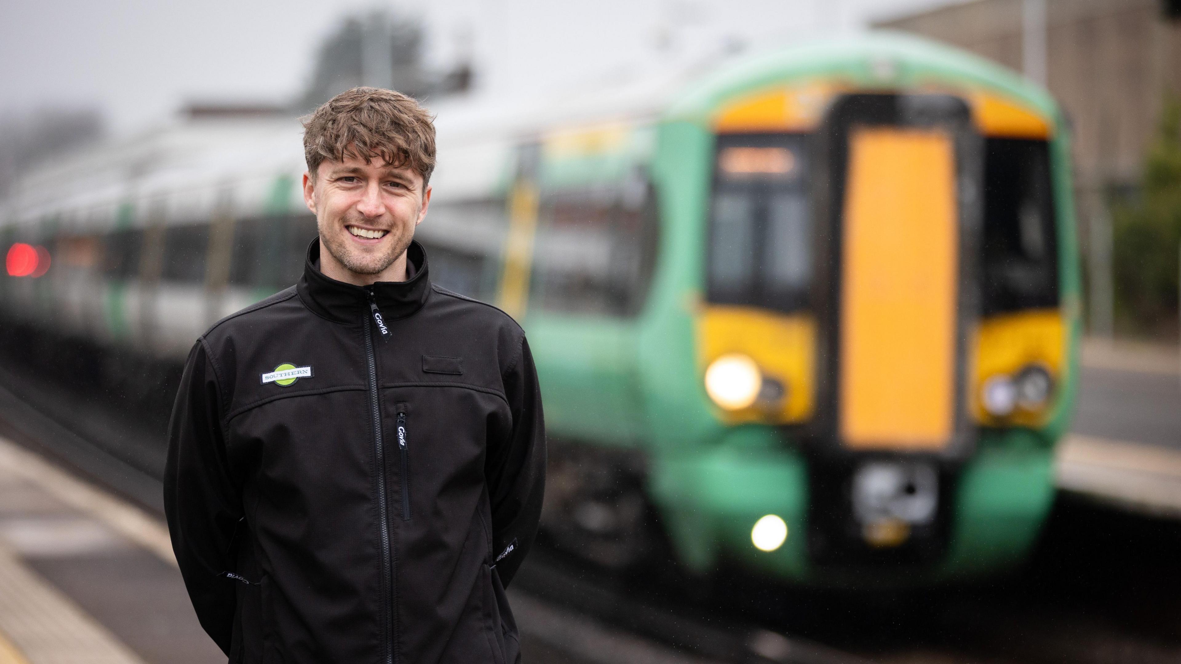 Jake Robinson standing on the platform wearing a black jacket with a green Southern Rail logo on the right. Jake has curly brown hair and a fringe down to his eye brows and is smiling at the camera. There is a green and yellow southern rail train behind him ready to depart from the station 