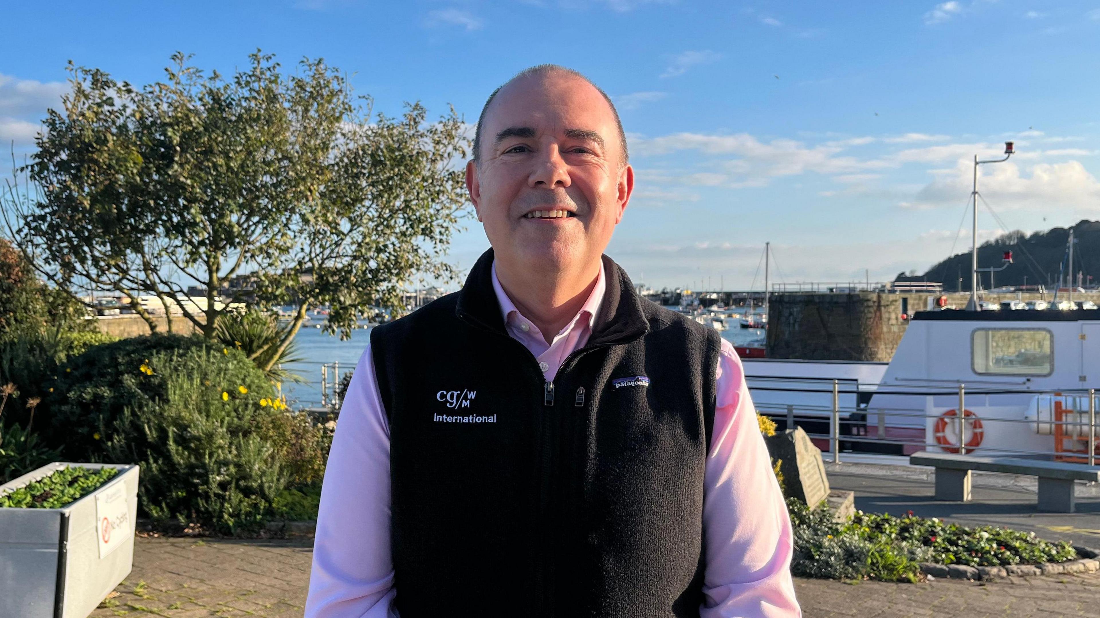 Andy Finch stood by Guernsey's harbour. He is wearing a pink shirt and a black gilet. He has dark balding hair. A white ferry can be seen in the background along with bushes and shrubs 