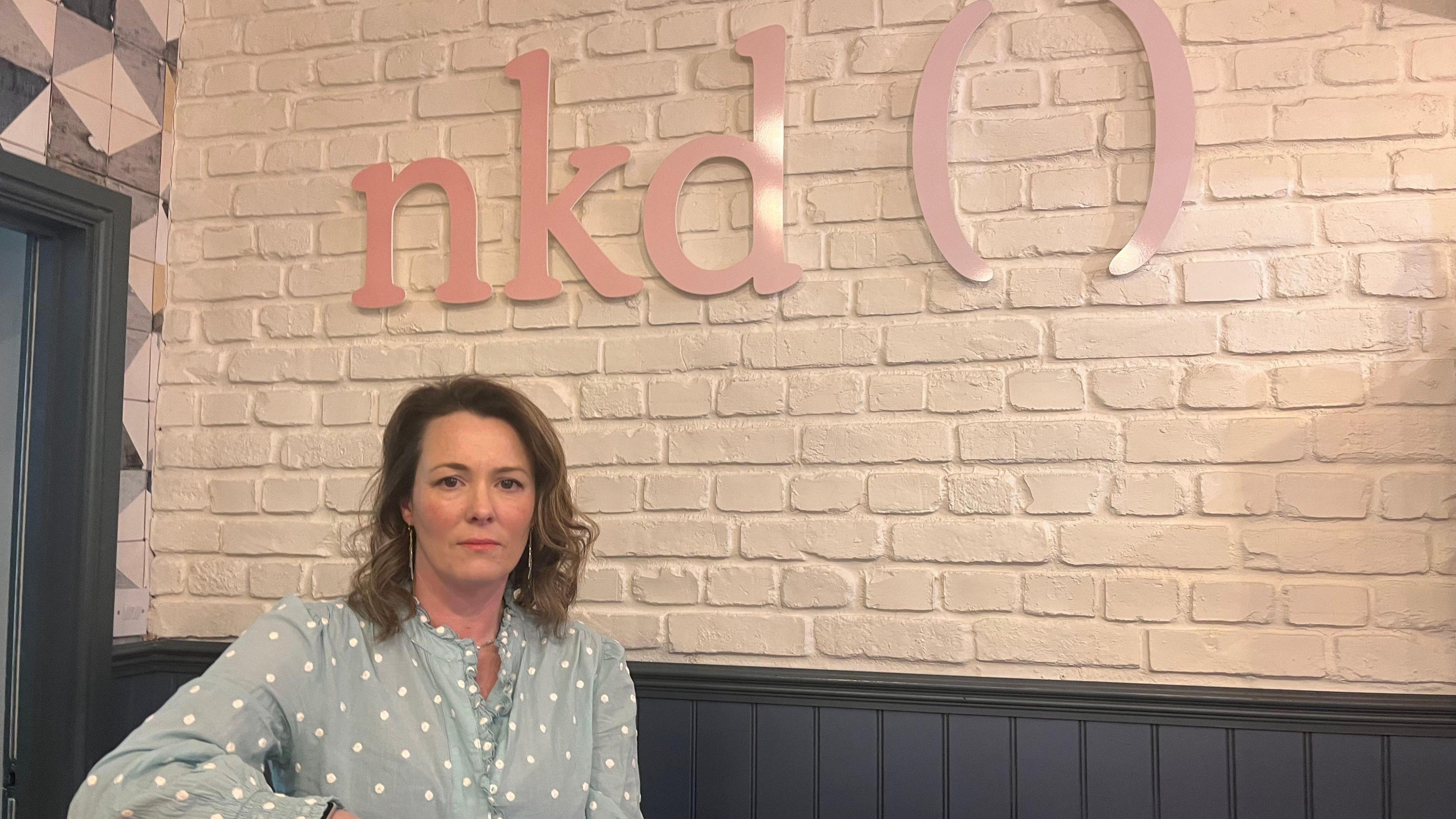 A woman with long dark hair, wearing a blue shirt with spots, standing in front of a salon sign saying nkd ().