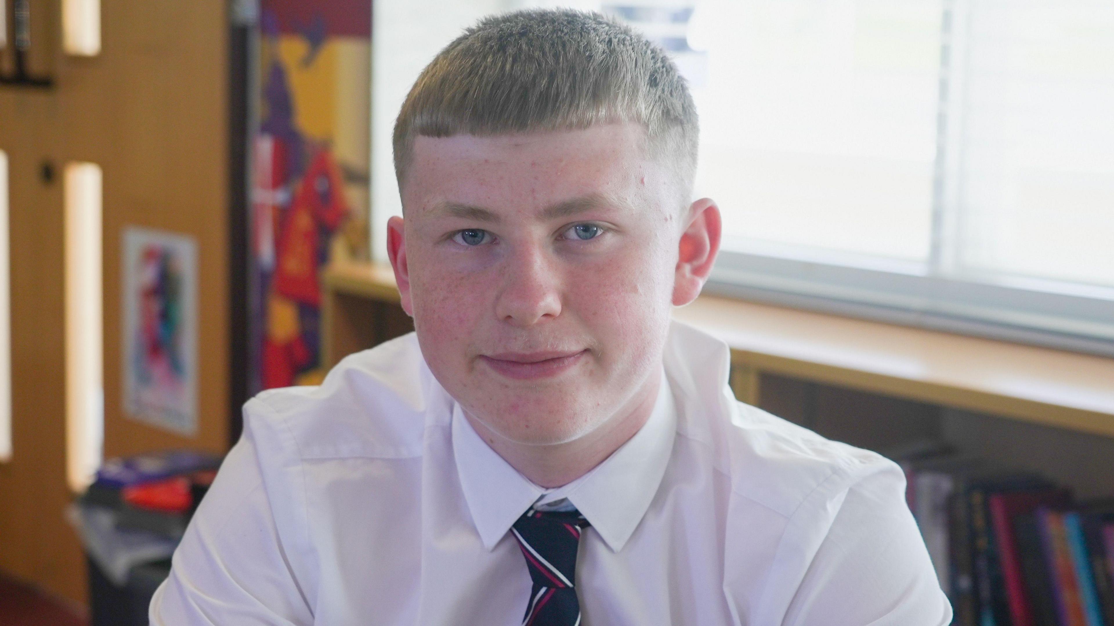 Bachgen yn ei arddegau gyda gwallt golau byr yn gwisgo crys gwyn a thei ysgol yn edrych ar y camera gyda silff lyfrau tu ol iddo