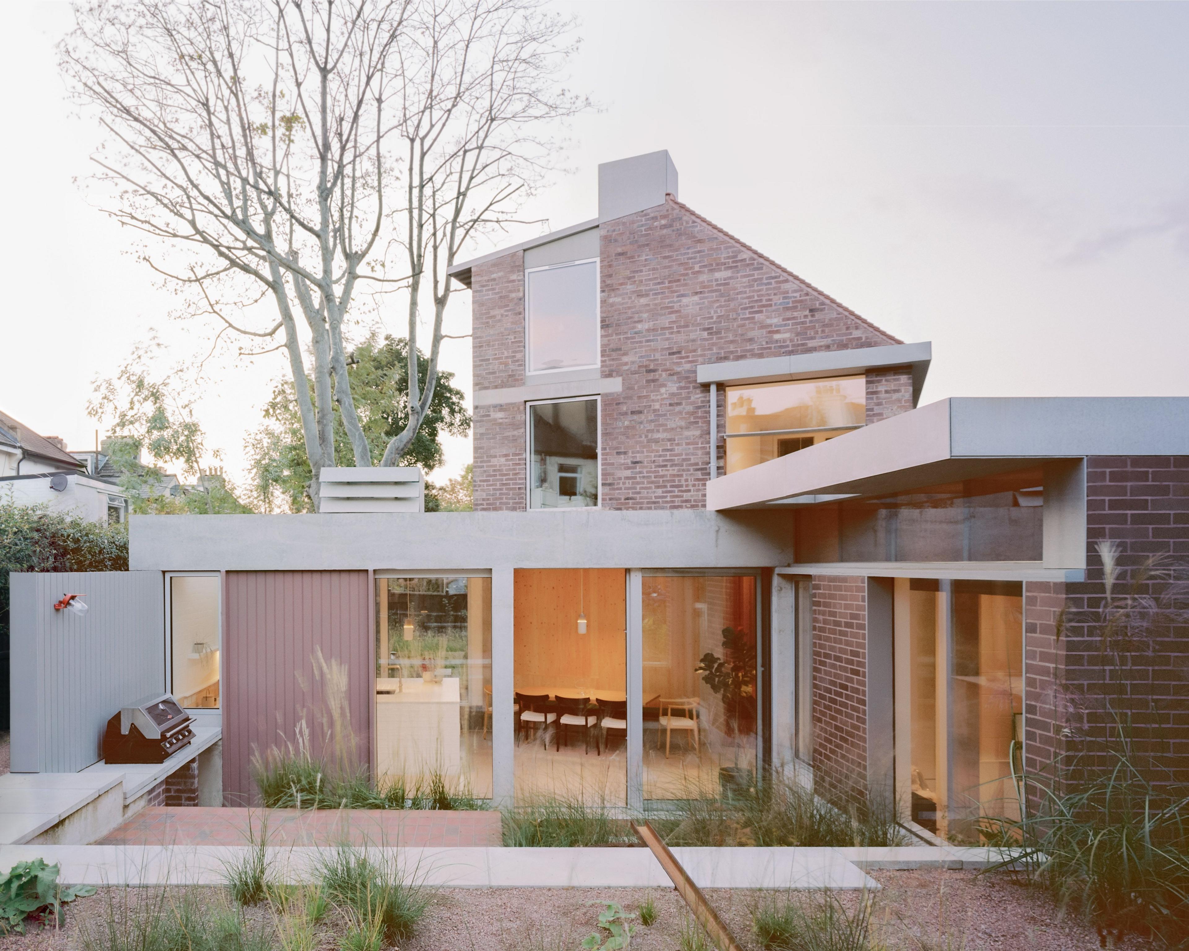 Back garden of Six Columns which shows tussocks of grasses planted randomly in gravelled areas. There is also a view into the inside of the house which has large floor to ceiling windows and doors on the ground floor and the first floor of the house with three windows.