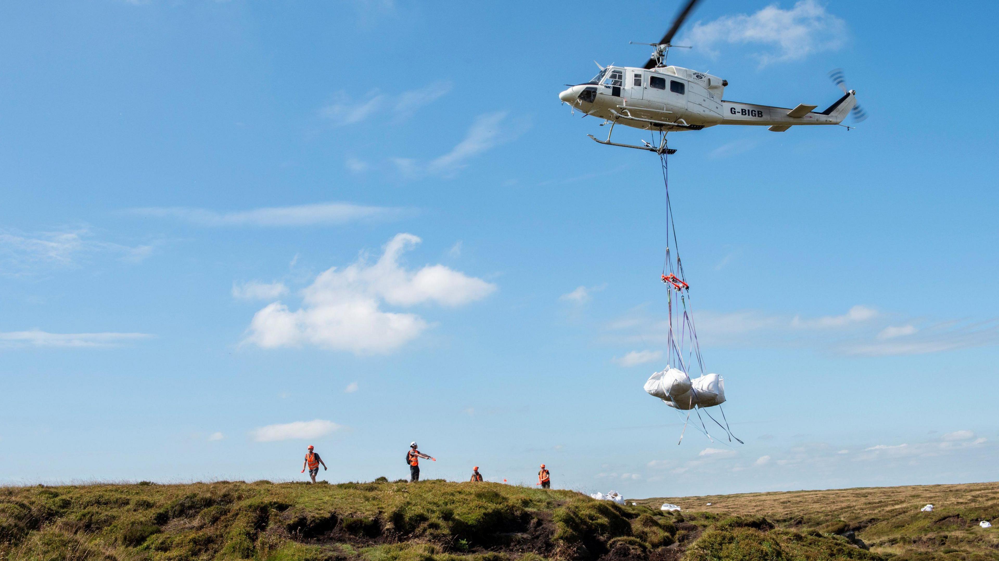 A helicopter carries white bags suspended on a line, marshalled by four people on the ground in hi-vis