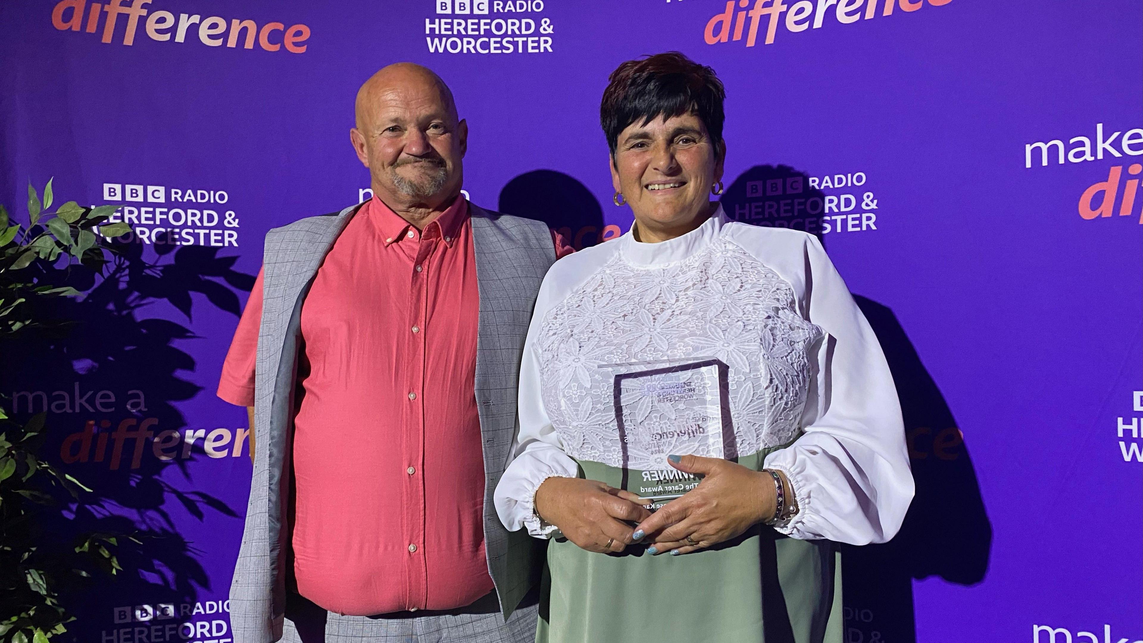 A man and a woman stand in front of a BBC backdrop. The women is holding an award. She is in a white blouse and green skirt. The man is in a pink shirt