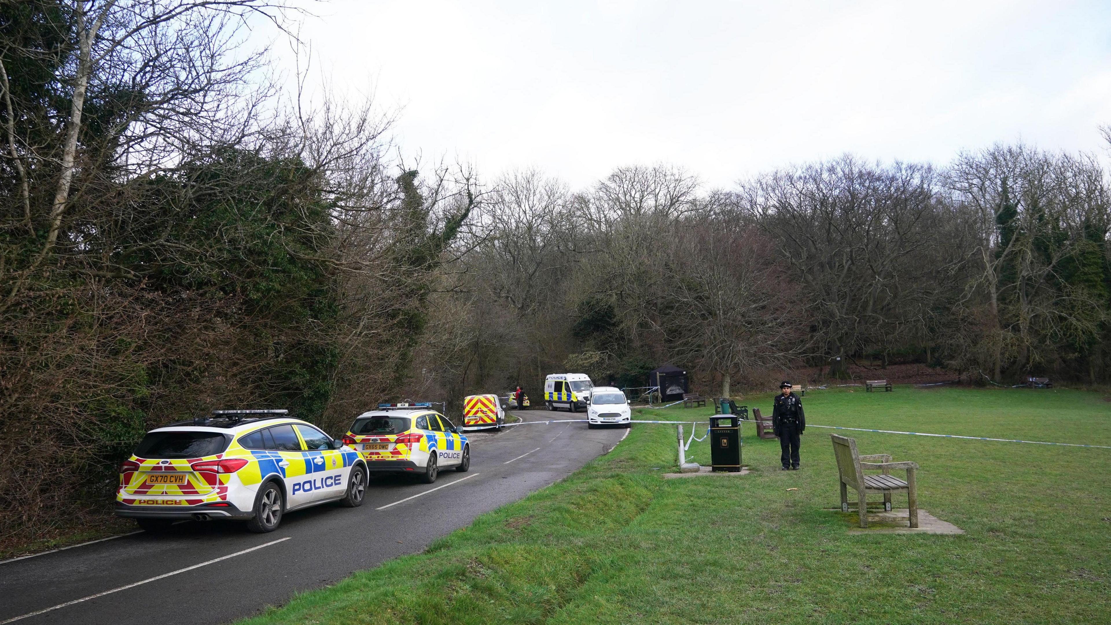 Police cars are seen parked along the side of the road next to a green space where there are park benches and trees that have no leaves on them. A lone police officer is seen facing the camera in front of some police tape.