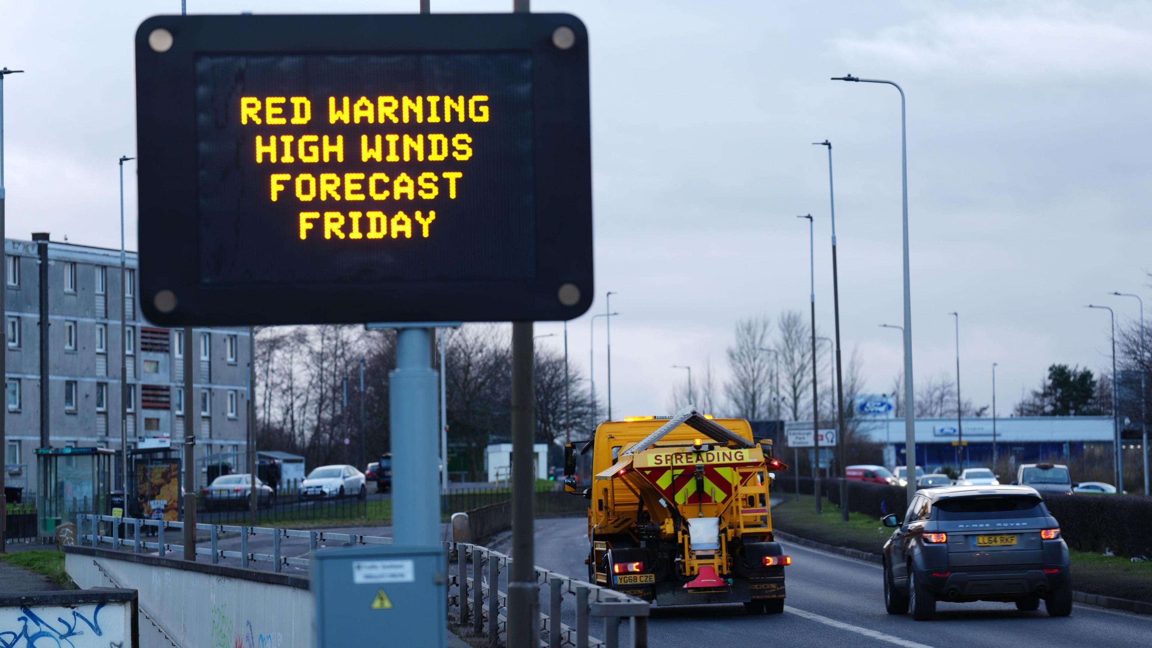 A black road sign displaying a red weather warning in orange letters for Friday on Calder Road, Edinburgh. A block of flats is on the left and a gritter passes on the road to the right.