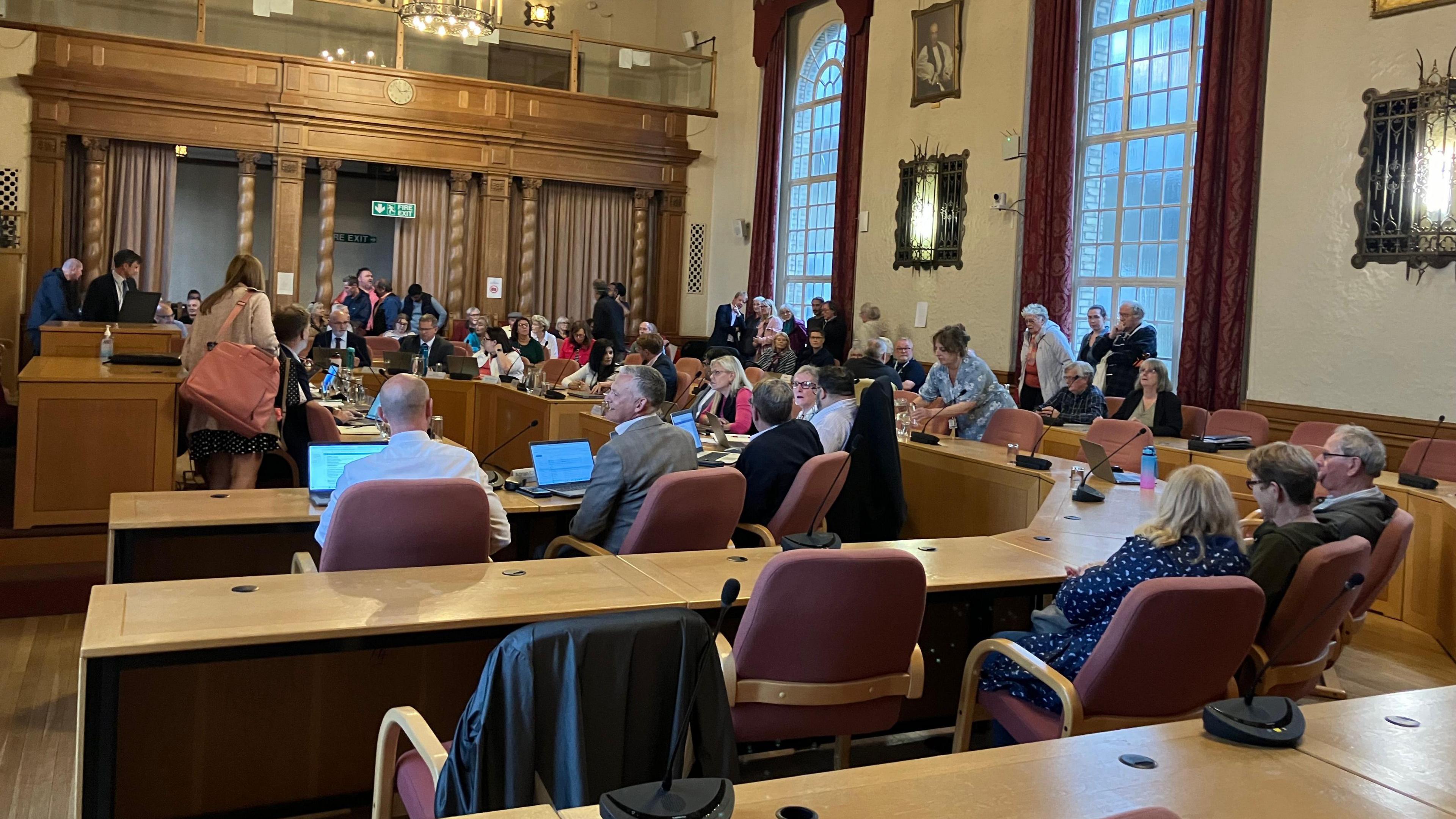 Inside a town hall chamber a group of people sit at desks in front of laptops and microphones