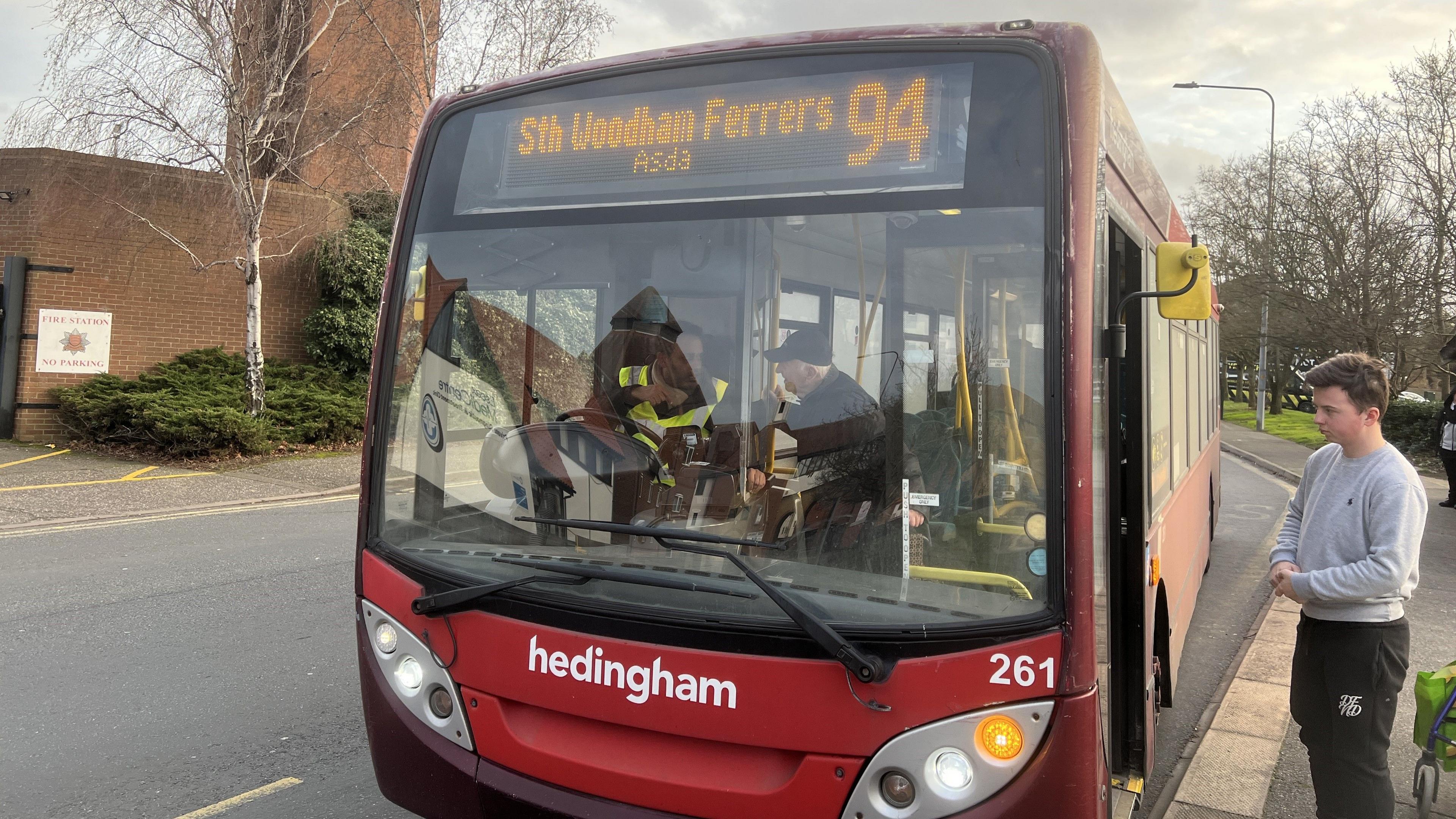A red single-decker bus at a bus stop. Its destination board reads: "South Woodham Ferrers Asda 94". A passenger waits to board.