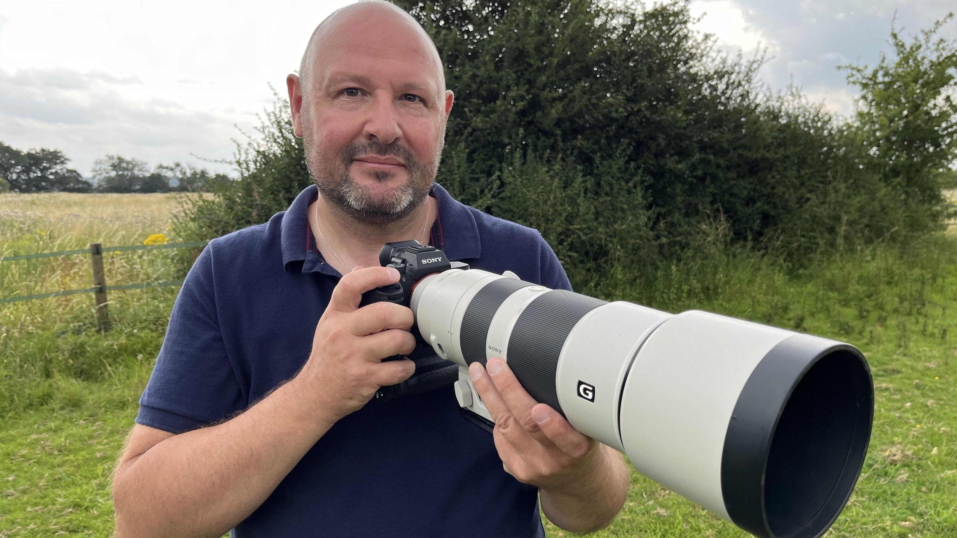 Colin Shead at the farm holding his camera and smiling at the camera. He is wearing a navy blue top