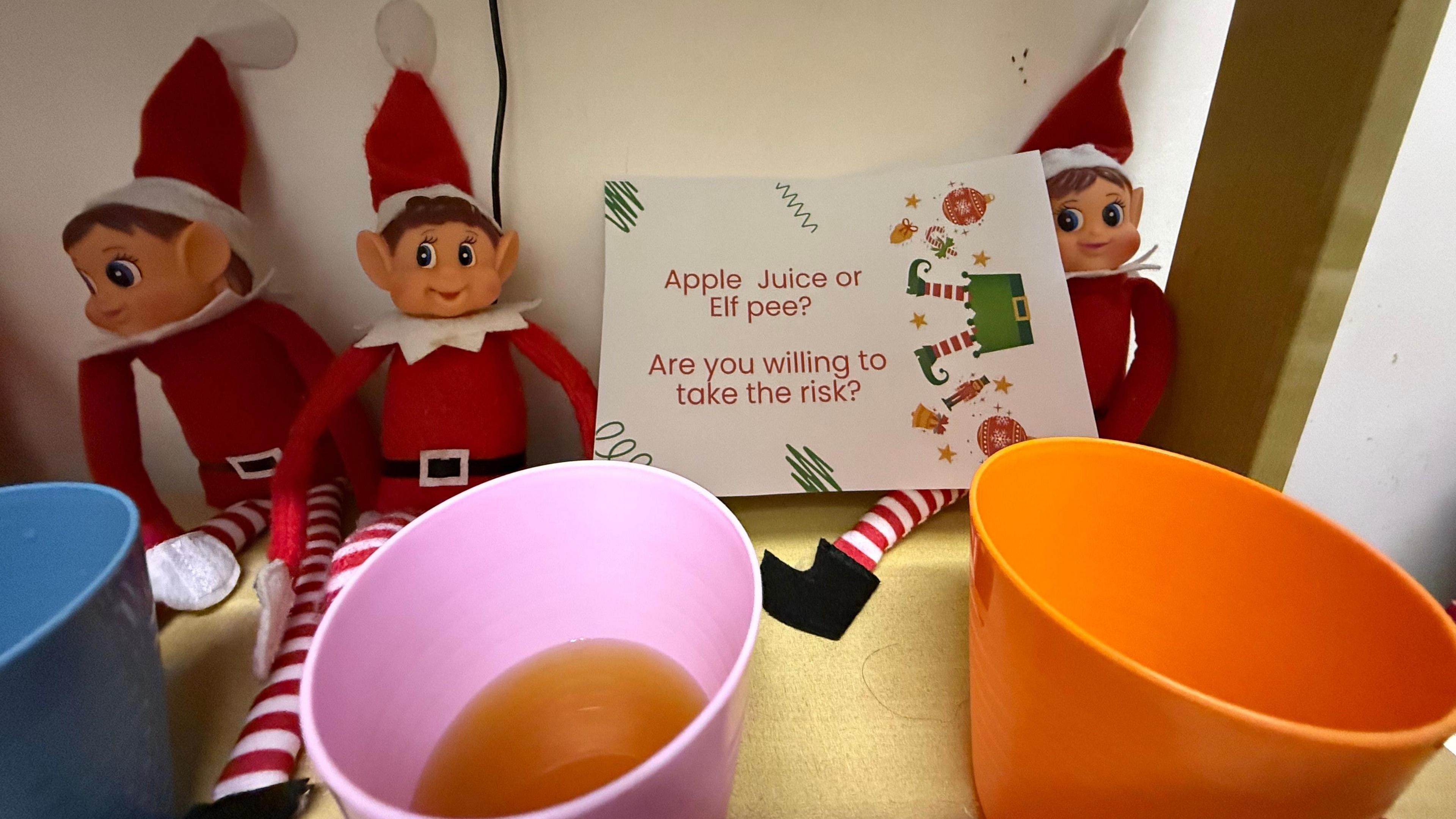 Three elves and three cups with apple juice in. One elf is holding a sign that says, "apple juice or elf pee? Are you willing to take the risk?".