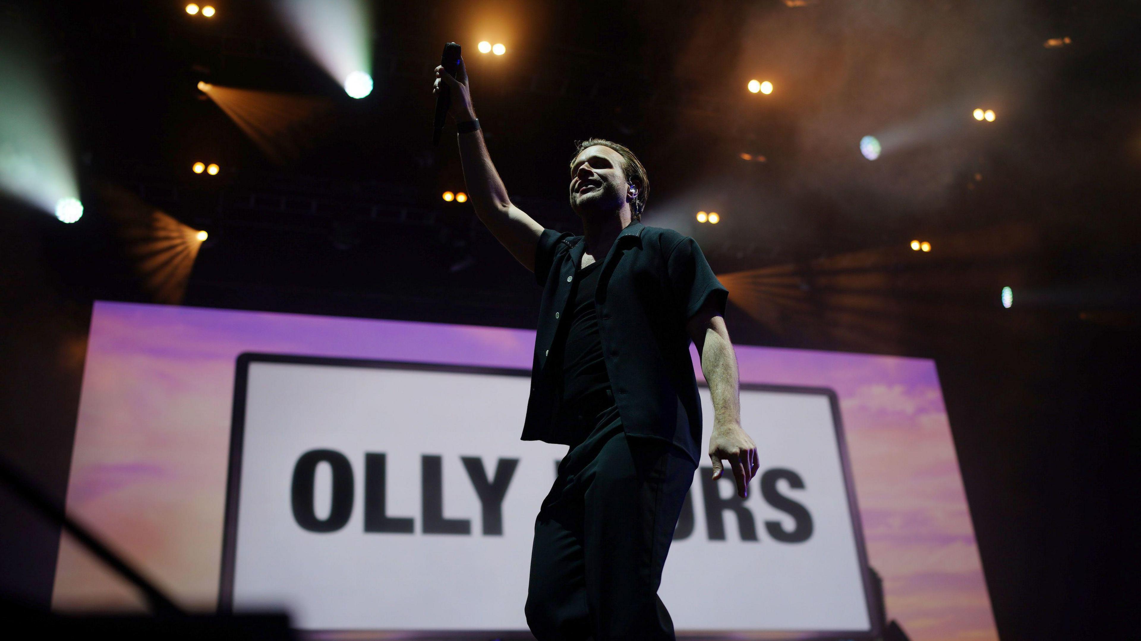 Singer Olly Murs on stage waving a microphone to the crowd, with his name in large letters on the screen behind