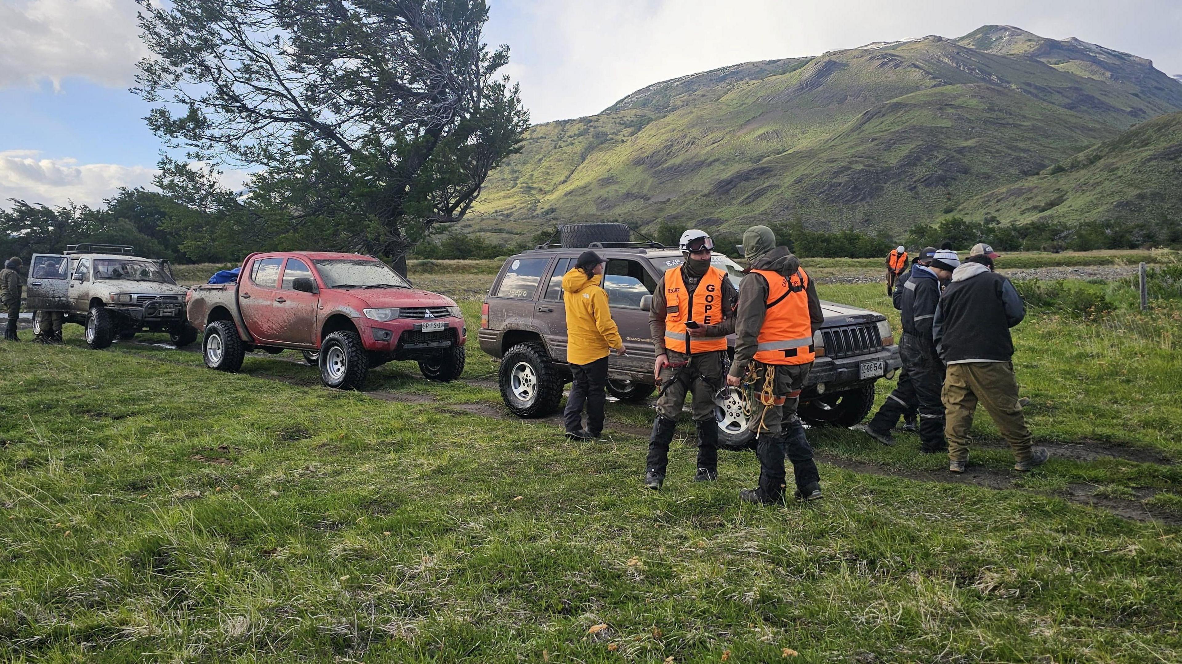 Three large vehicles parked on a grassy field. A group of people are standing next to the cars. They are wearing hi-vis safety gear. A rocky hillside is in the distance behind them.