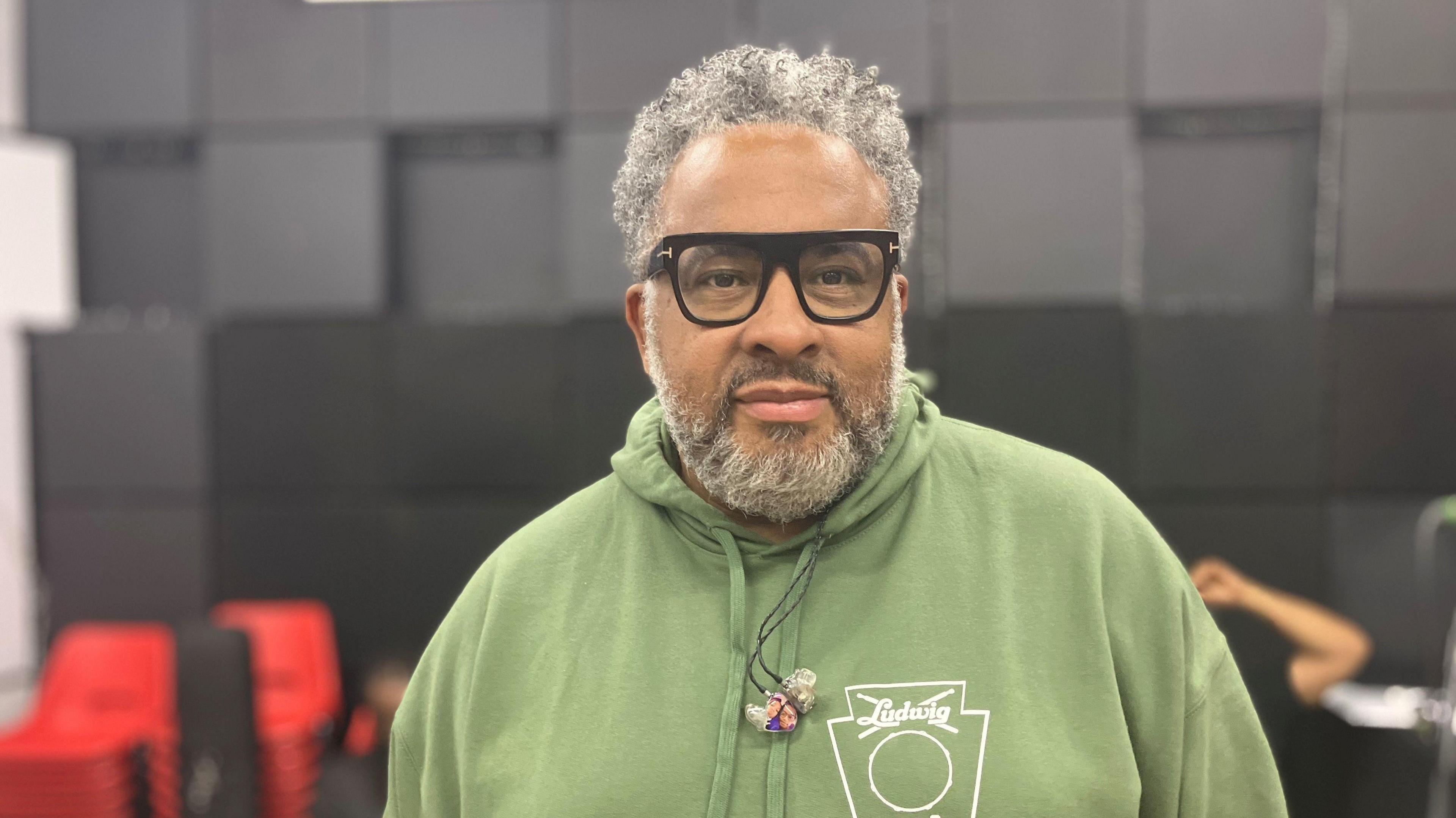 Donovan has green hooded top on and glasses. He has grey hair and a beard and is inside the recording studio.