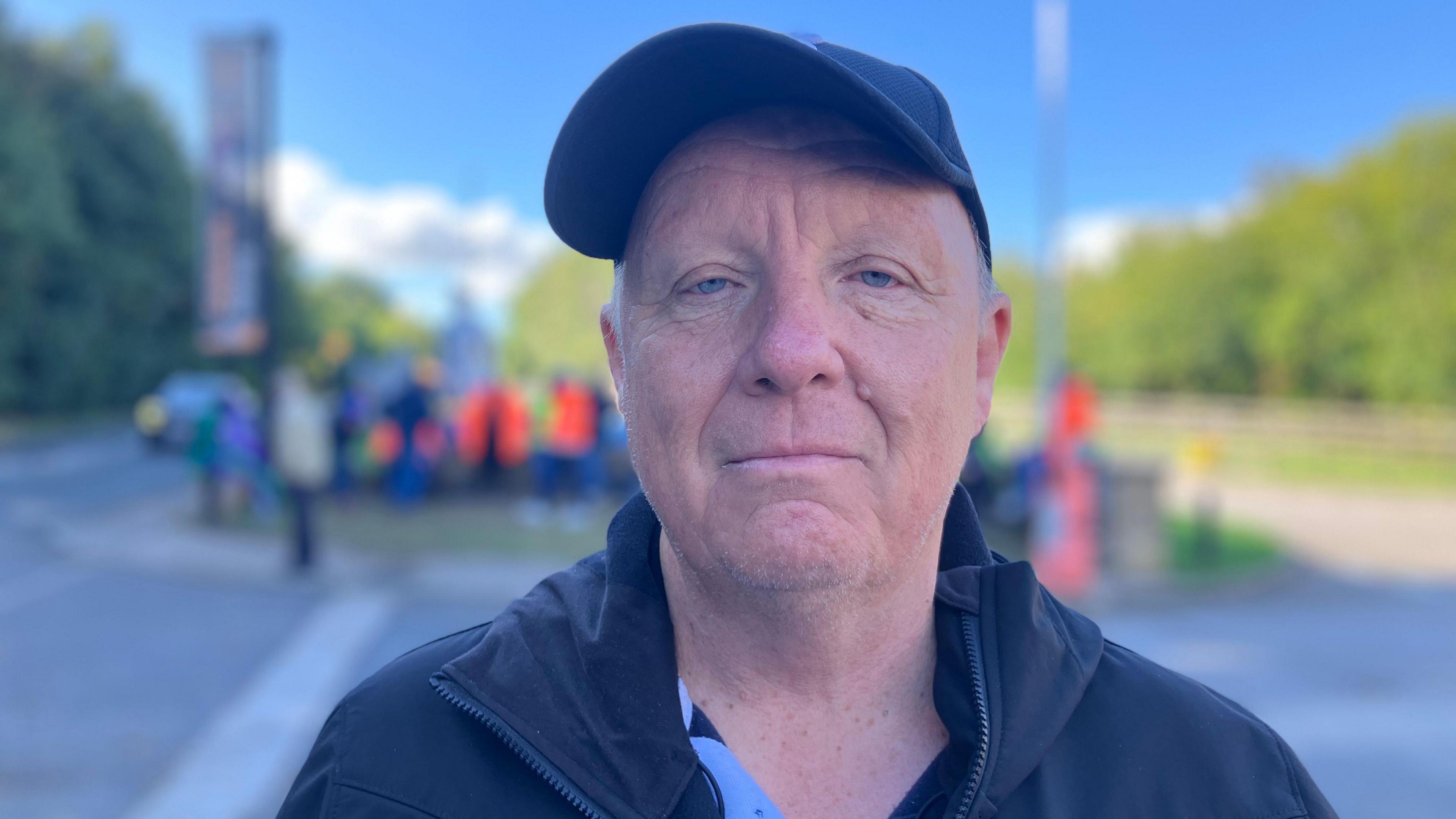 A man wears a dark cap and looks directly at the camera. He wears a black coat, behind him, picketers are blurred.