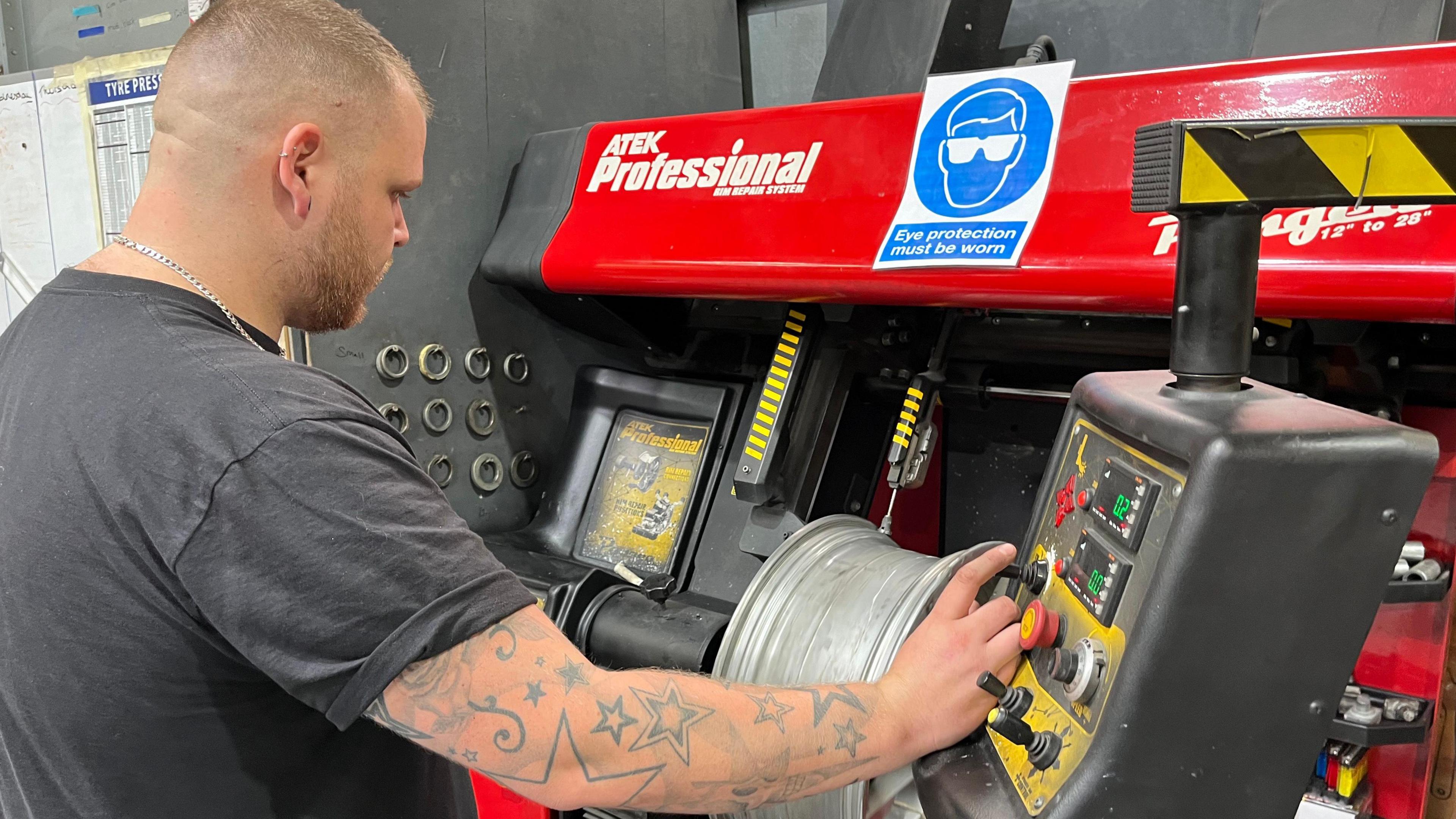 David Linforth stands in front of a machine that fixes buckled alloy wheels. He has his back to the camera and faces the machine, he is wearing a grey T-shirt and silver chain. His arm is outstretched towards the control board. 