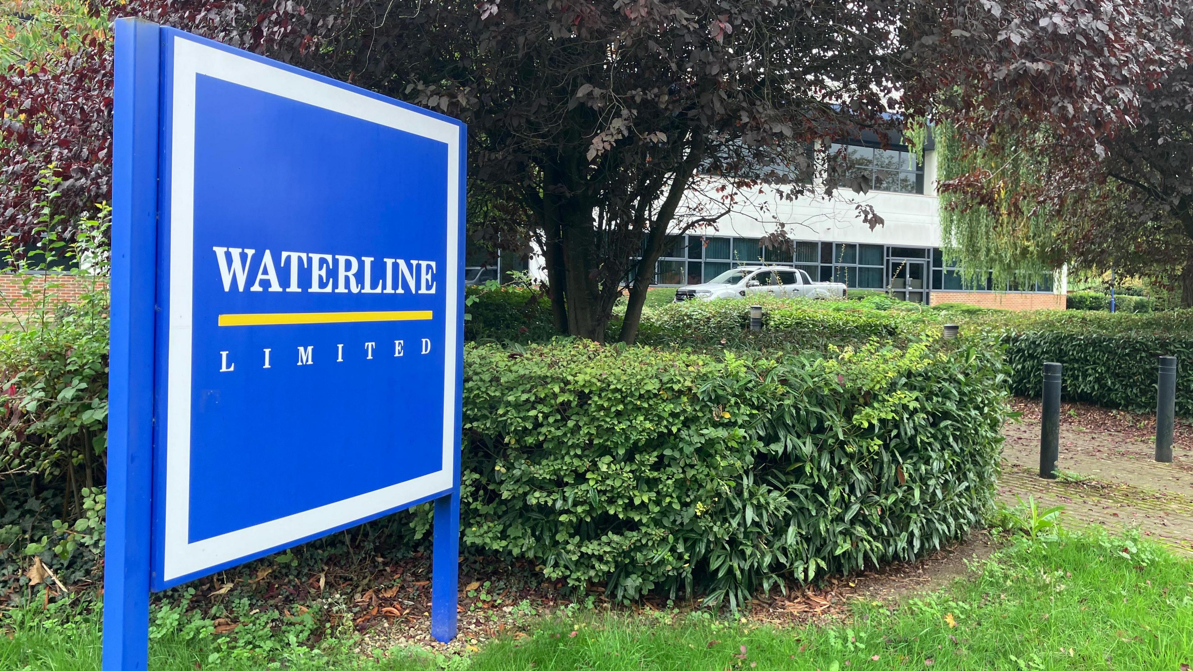 There is a large blue sign with a white border that takes up much of the left hand side of the picture. It says "WATERLINE" on it with a yellow line underneath it. Under that it says "LIMITED". Around the sign is a green hedge and behind that trees which appear to have purple leaves on them. Behind that a large white office-style building can be seen with large windows. A silver pick-up truck can be seen parked in front of the building.