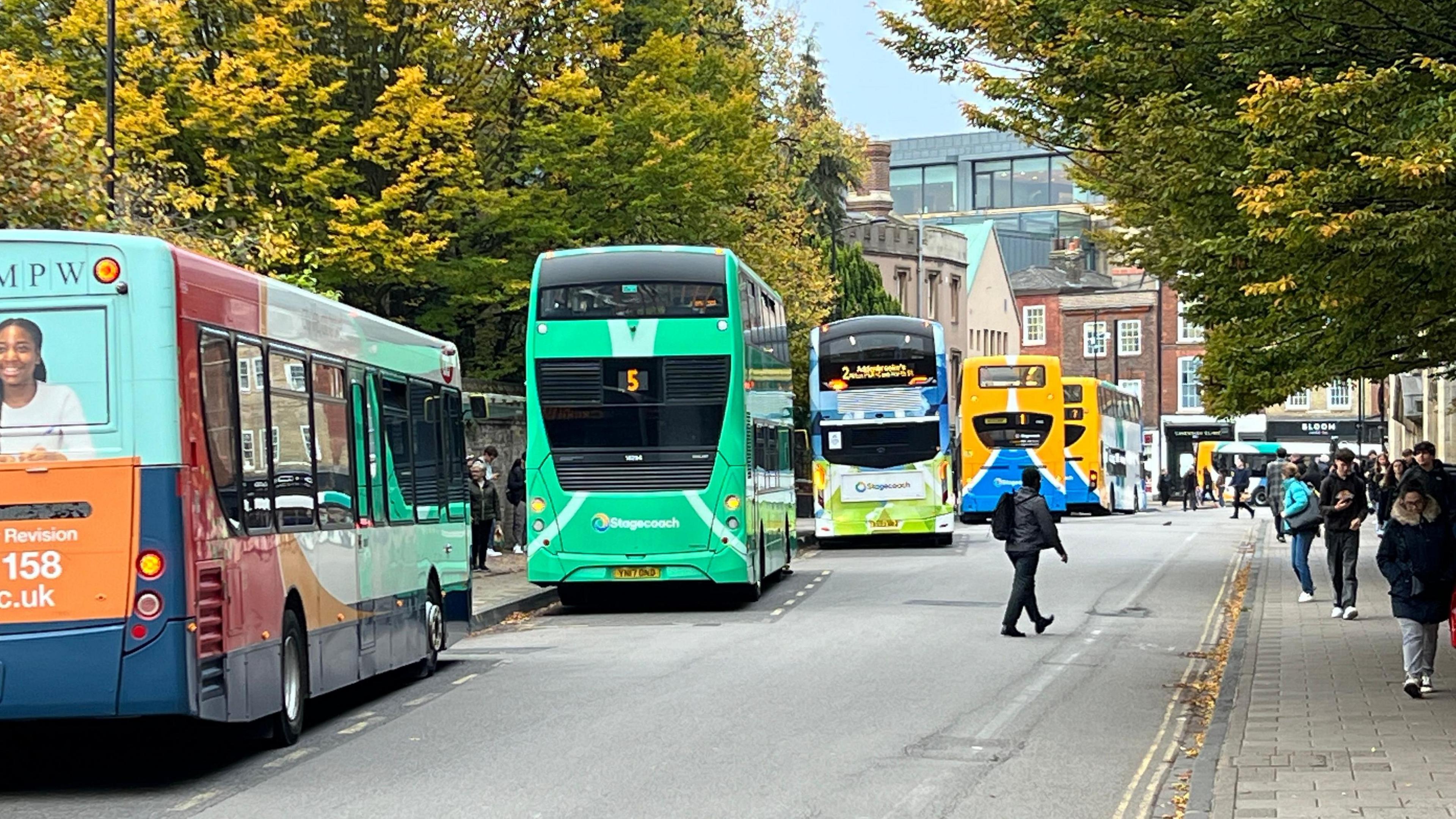 A street with five parked buses on the left hand side. To the right is a path with lots of pedestrians.  