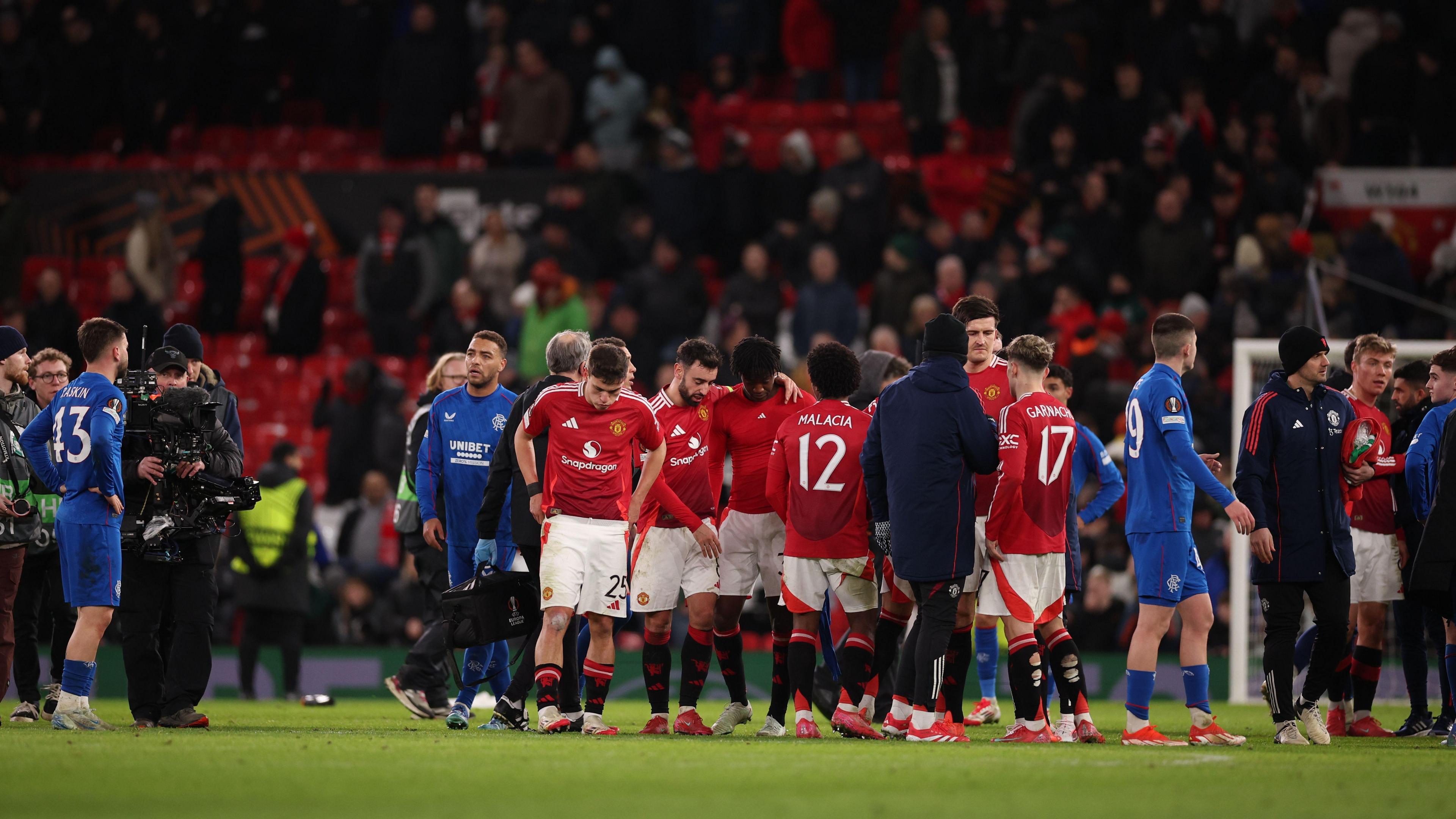 Manchester United and Rangers players on the Old Trafford pitch after the match - the Rangers players, in all blue kits, look dejected with hands on hips, while the Manchester United players - in red shirts and white shorts look tired, some with their arms around each other