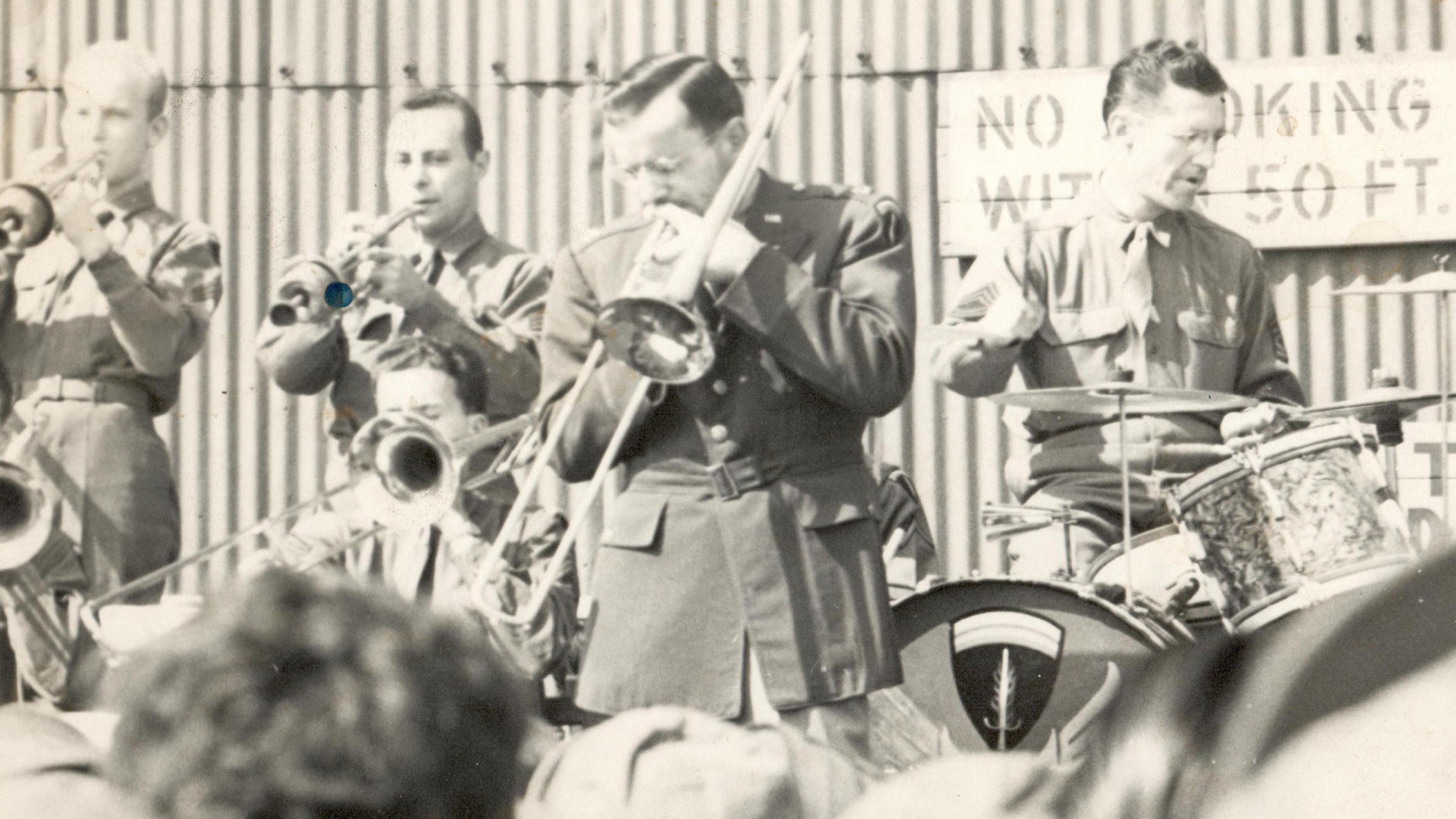 A black and white image of the band, showing four musicians, all wearing military uniform. Drummer Ray McKinley is on the right with a small drumkit. To his left is Glenn Miller on the trombone.
