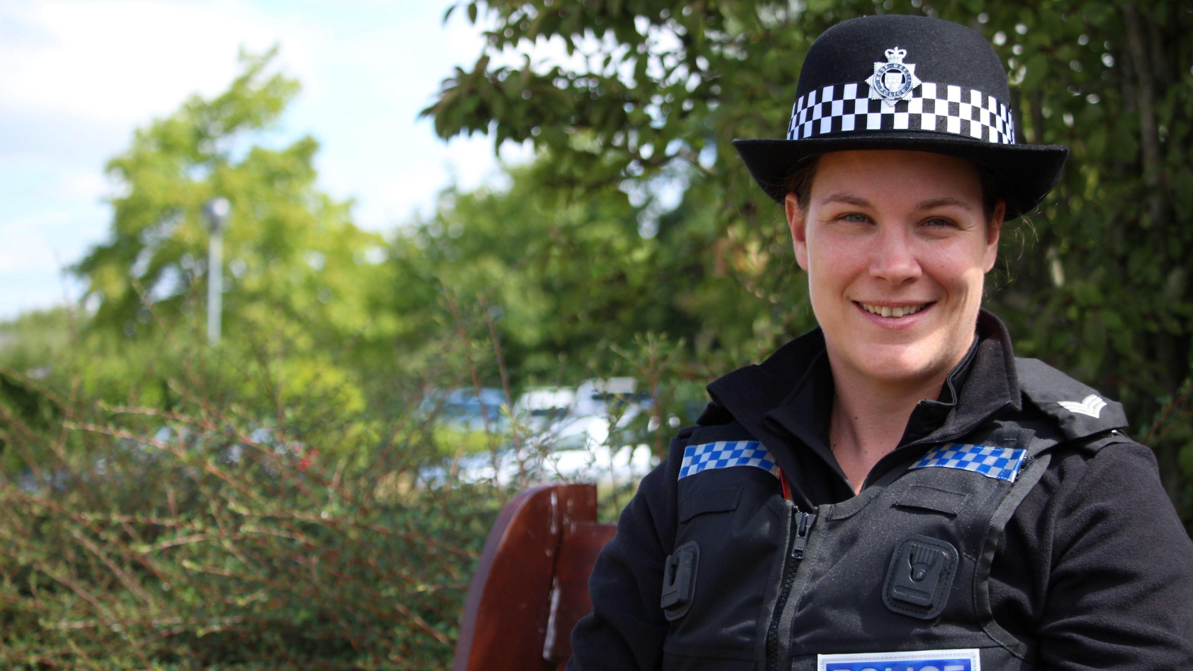 Sgt Kat Scrimshaw is in a black vest and black long-sleeved top. It has two stripes of blue and white checkerboard pattern. She is wearing a blue badge that reads "police". She is wearing a black police hat with a black and white checkerboard stripe, and the West Mercia Police badge in the centre. Behind her are green trees and hedges.