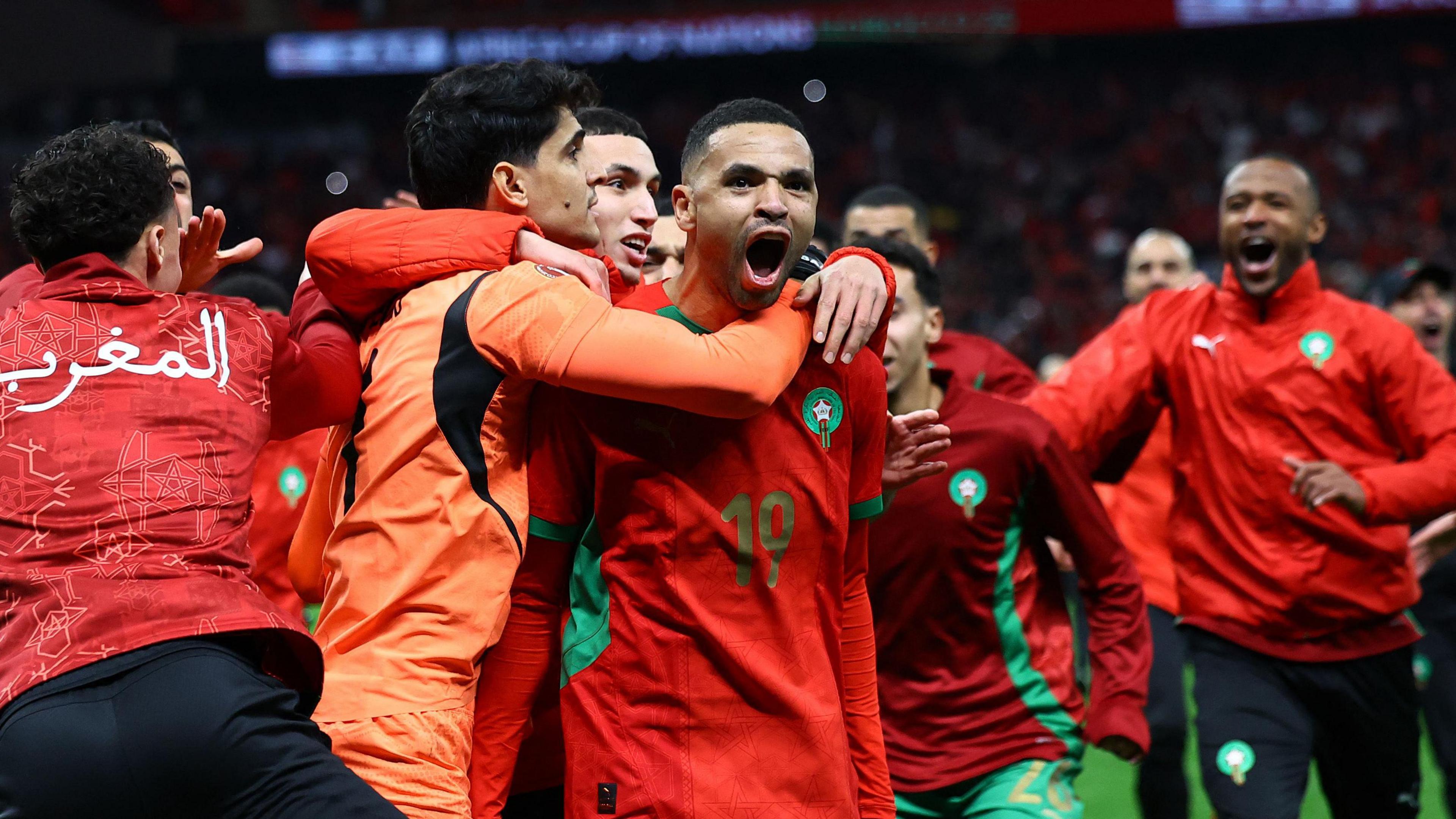 Morocco players and staff mob Youssef En-Nesyri after winning a penalty shootout against Nigeria at Afcon 2025. En-Nesyri seen from the waist up is in his red Morocco jersey and is opening his mouth wide to shout, while two players, including goalkeeper Yassine Bounou in orange, wrap their arms around him from behind