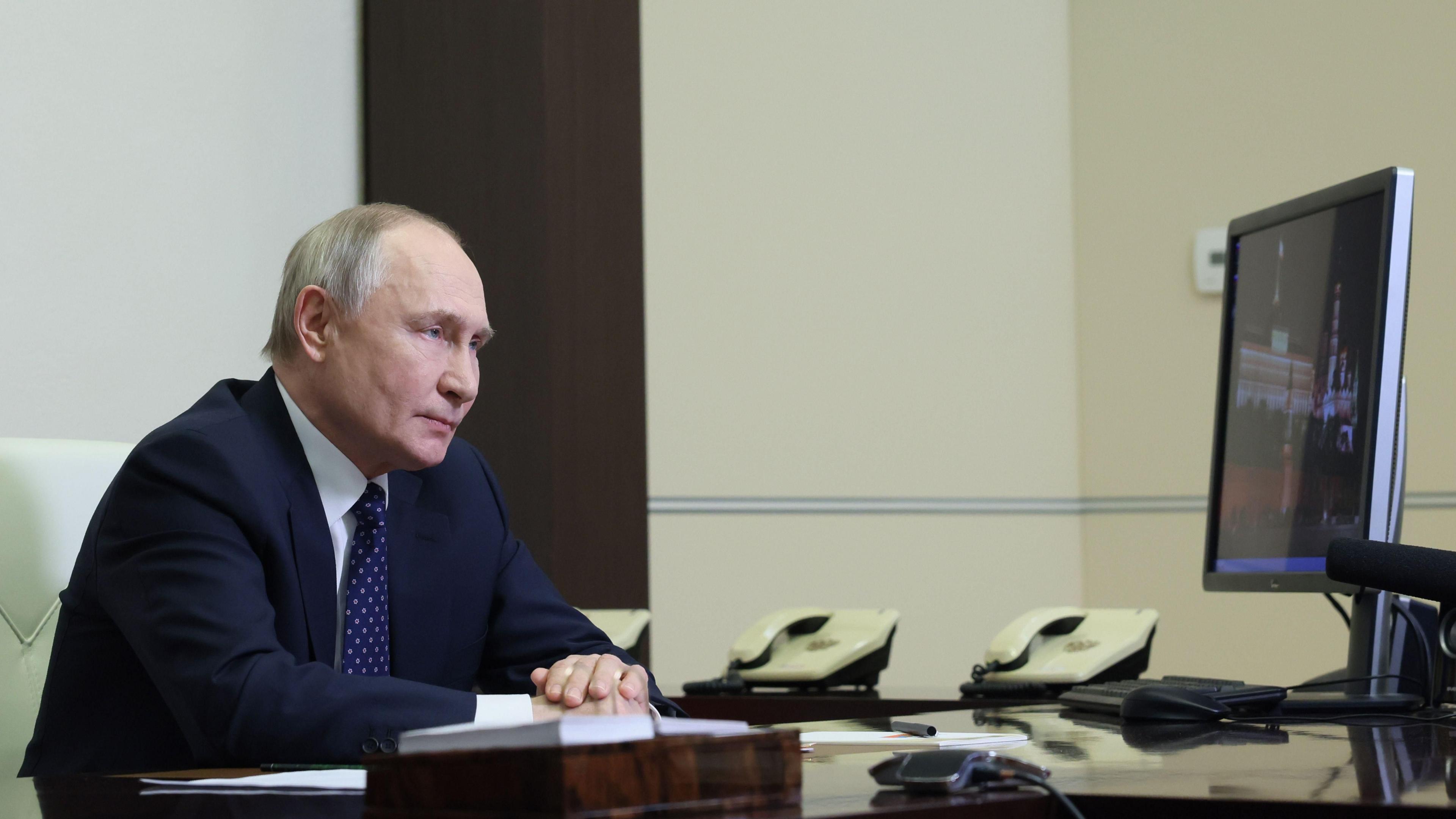 Vladimir Putin sits left of picture in profile, hands rested on his desk, looking at a computer screen on the right of the picture, as he chairs a video conference with members of Russia's Security Council.