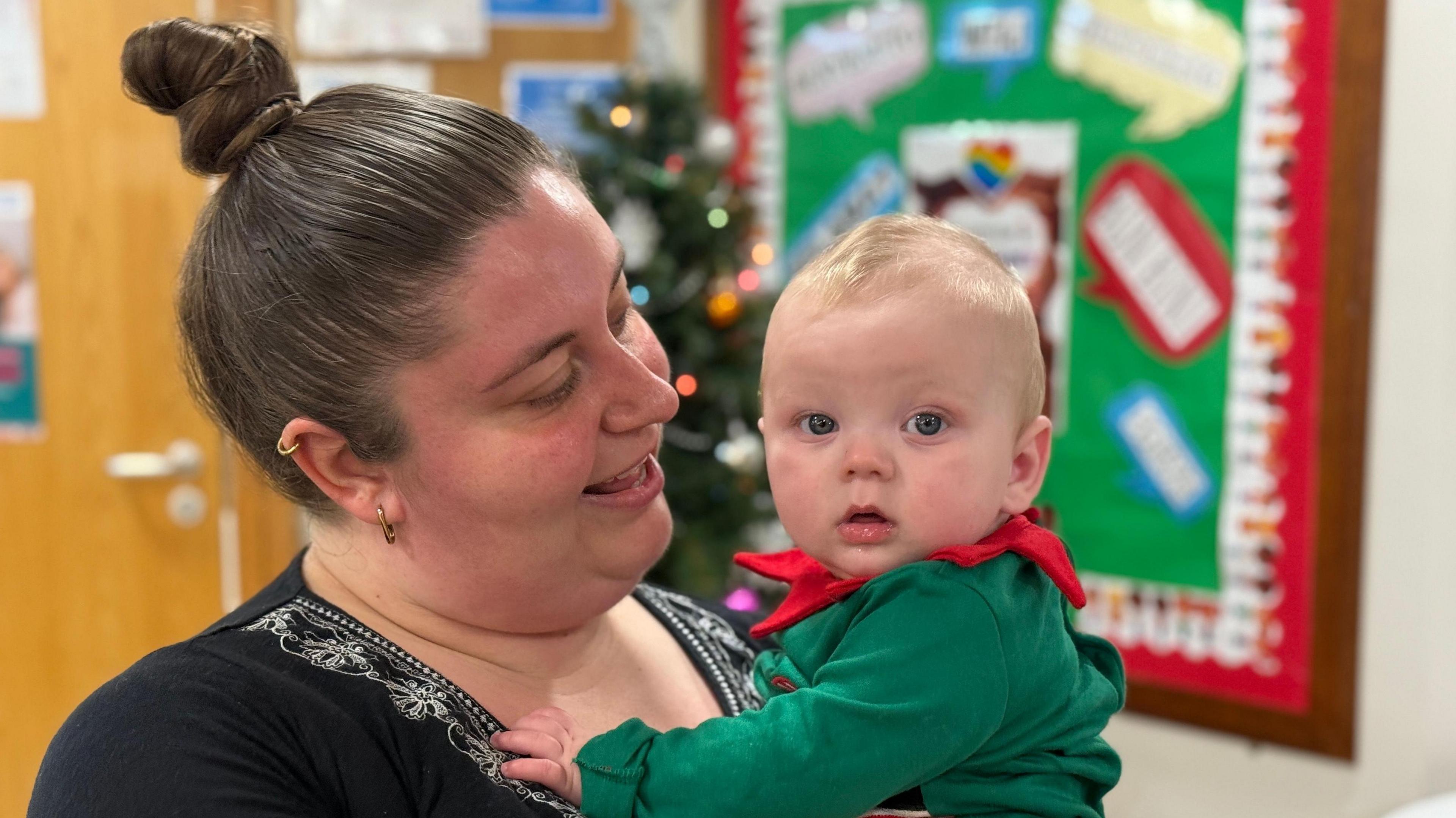 Rebecca holding a child. She is in a black top and has her long hair tied in a bun. The child is dressed in a green and red Christmas elf outfit.