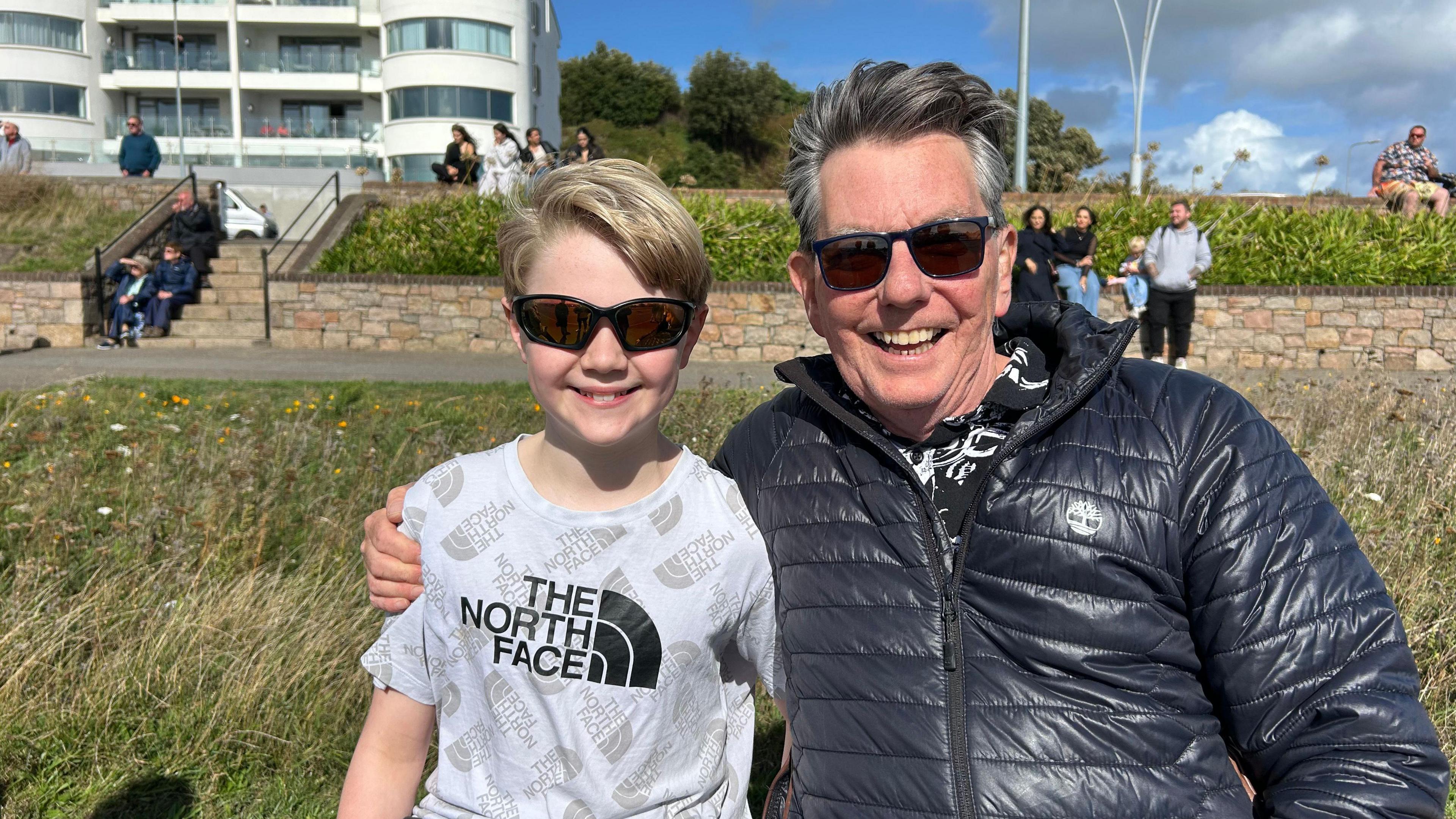 A smiling, grey-haired man in sunglass next to a blonde boy. The man hugs the boy's shoulder.