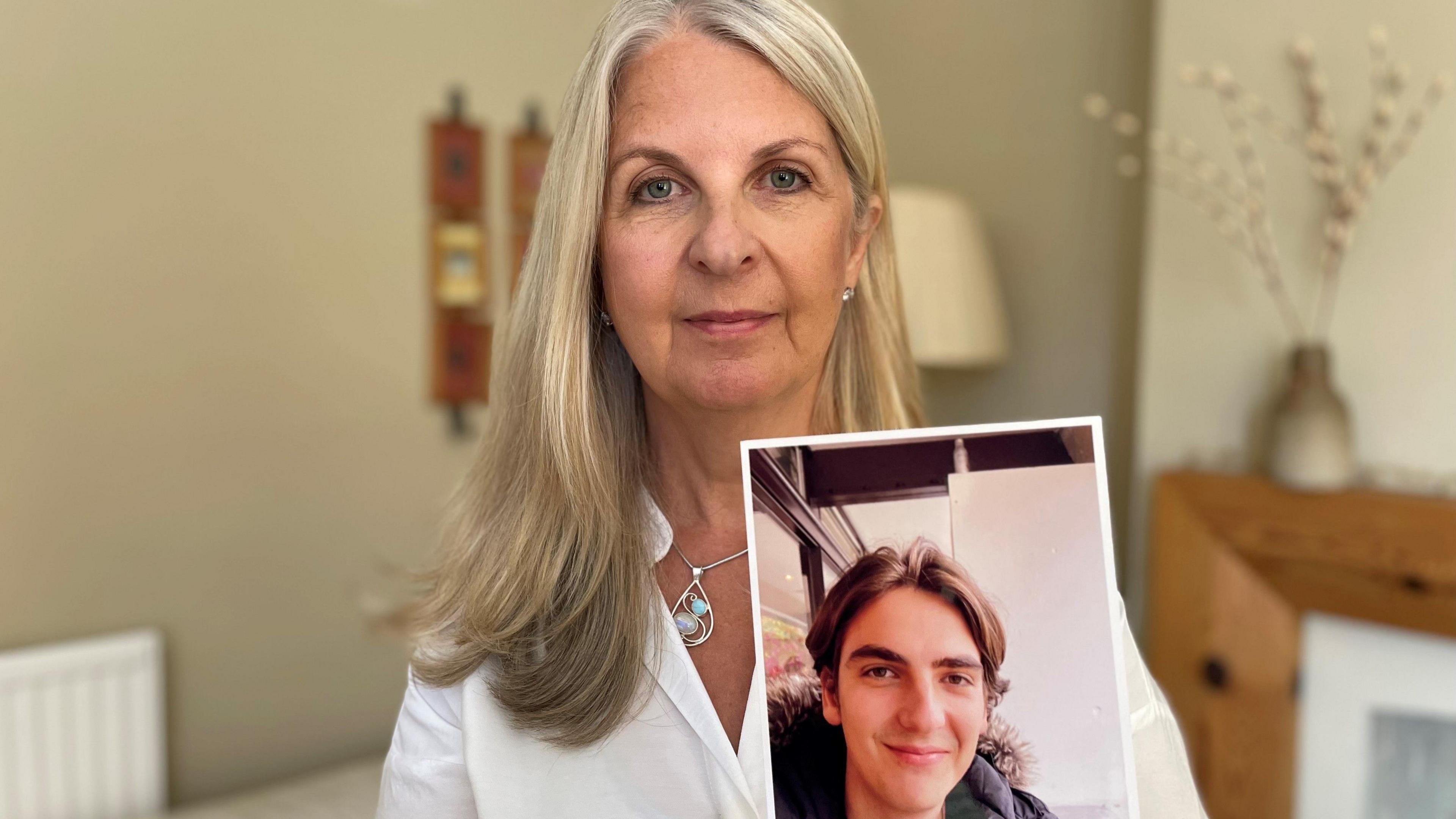 Anne Jacques faces the camera, wearing a white blouse, a silver pear-shaped necklace and silver earrings in her home. Her long, blonde hair is parted to the right. In her right hand she holds up a photo of her late son Alex Harpum. In the photo, Alex is smiling at the camera with his mouth closed. He has centre-parted brown hair, brown eyebrows and brown eyes. He is wearing a metallic grey puffer jacket with a hood.