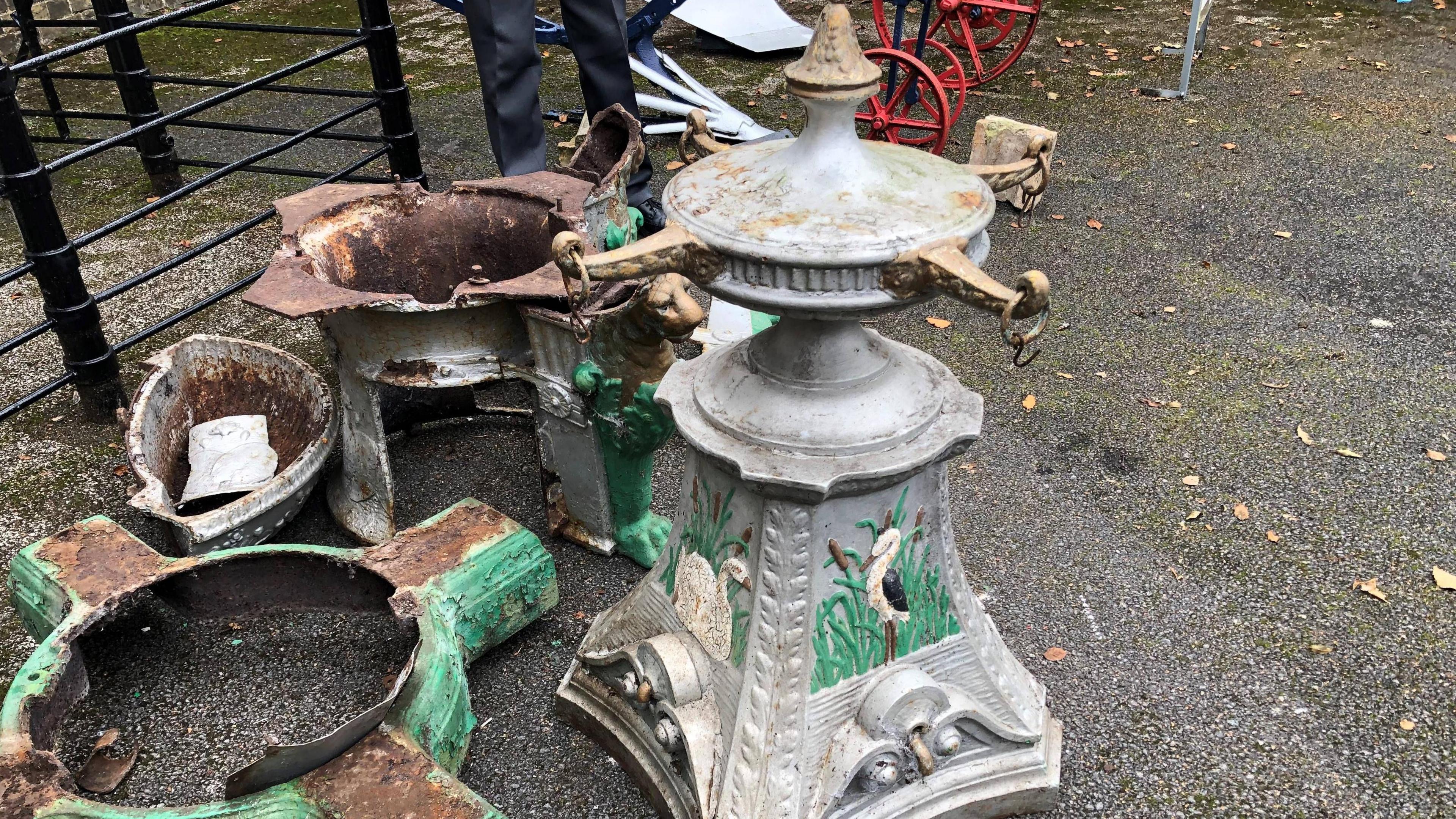 A heavy cast iron plinth painted silver with depictions of a swan on one side and a black and white water bird on the other. Both have reeds and bull rushes in the background, the object stands about 1m (3ft) high and has four gold arms, about 15cm (6 inches) in length coming off a circular shaped piece. Rings are attached to each arm, which would have originally had a chain on attached to a cup to collect drinking water. The large base, which is damaged, rusted and painted green, is next to it.