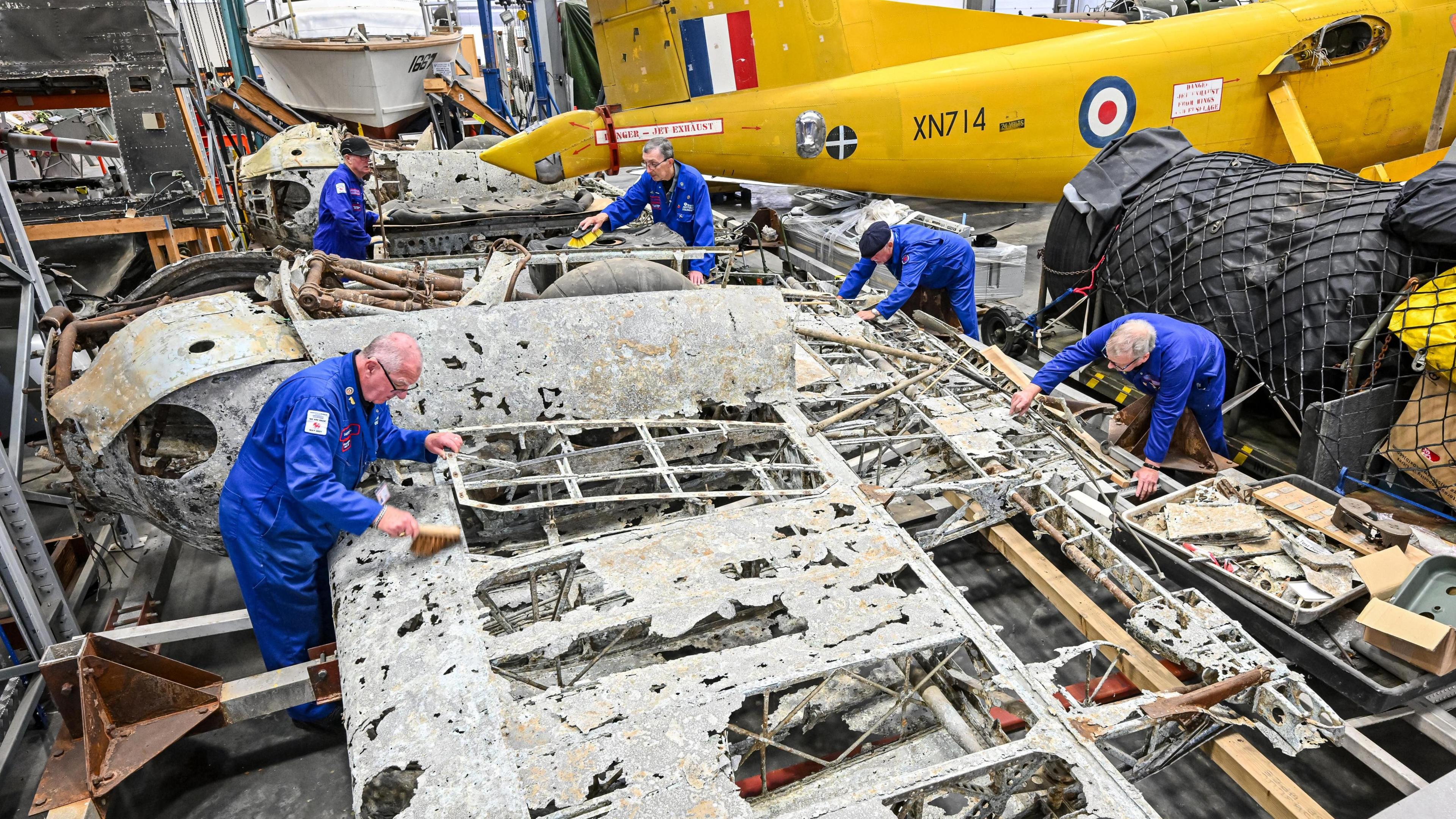 Five volunteers in dark blue overalls are working on the shell of a war plane. It is grey and metal, with holes across its wings. One of the volunteers is brushing the metal with a brush. 