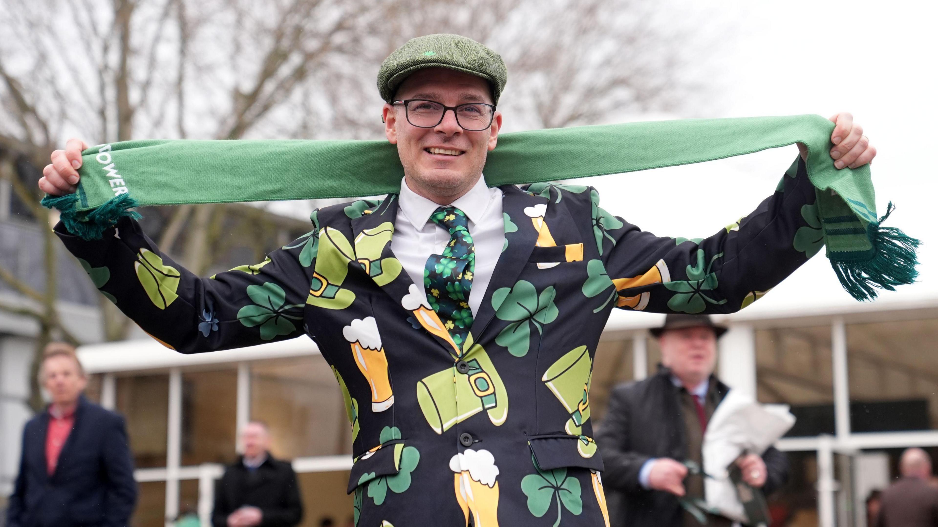A racing fan at Cheltenham 2026 poses holding up a light green scarf. He is wearing a jack covered in Irish logos such as shamrocks and leprechaun hats. He is also wearing a light green cap