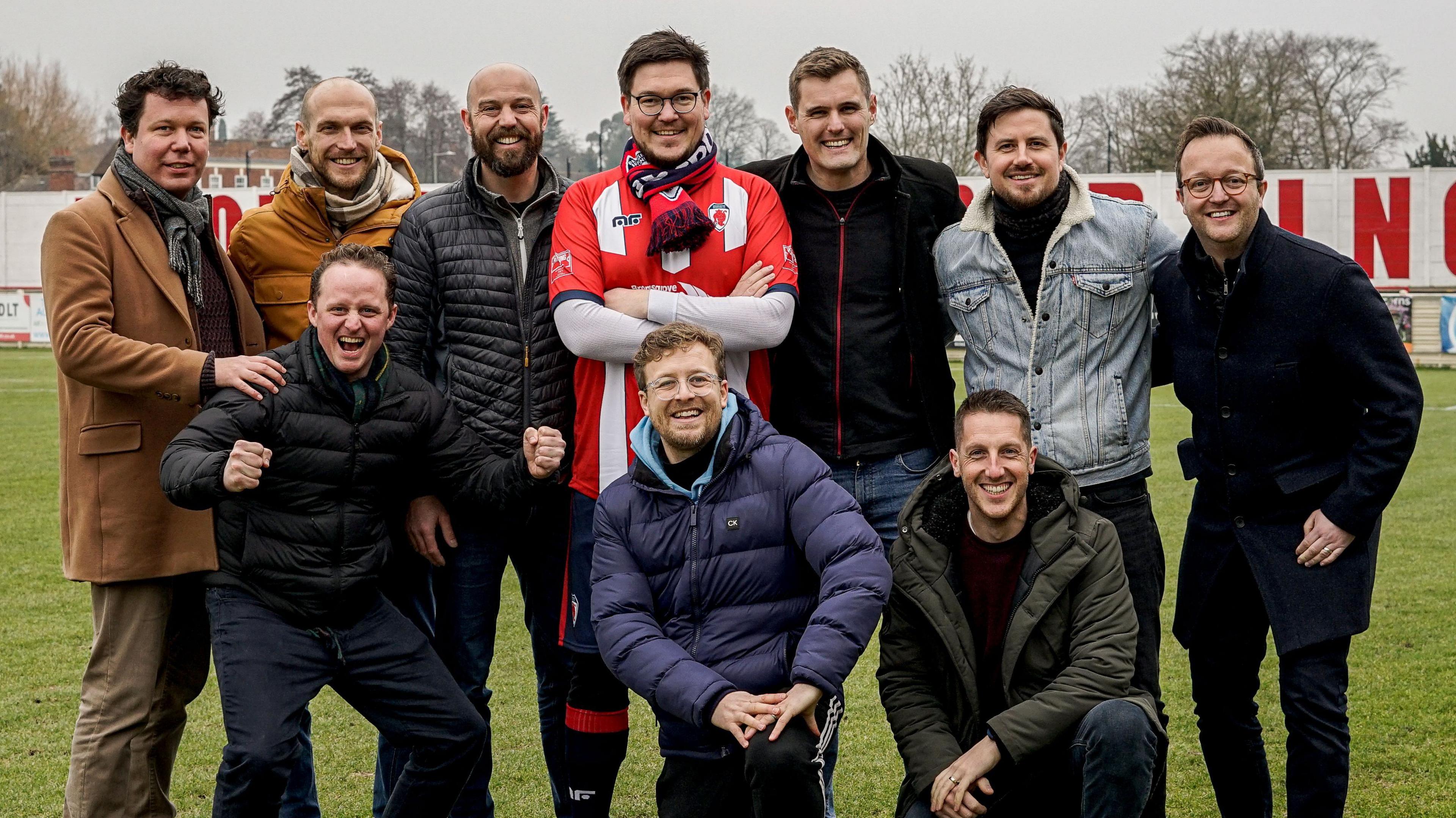 A group of men standing together on a football pitch. Two men are on one knee at the front, with eight others around them in a semicircle. The sky is grey in the background and the top of a few trees and buildings can be seen. The men are all wearing winter coats and dark trousers, aside from the man in the centre who has his arms crossed and is wearing a red and white striped football shirt. 