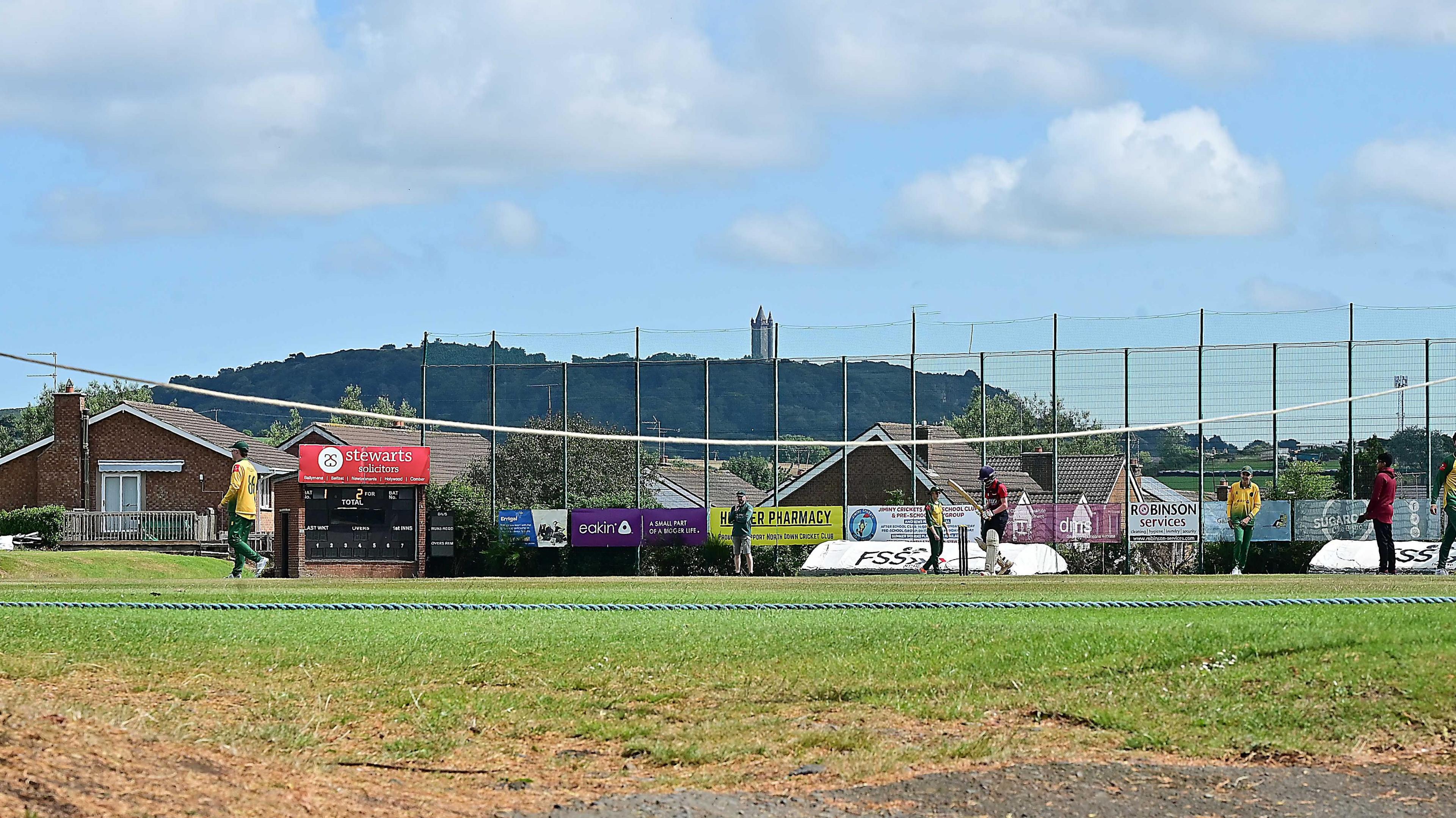 A cricket game being played. Nine layers are on the pitch, some in green and yellow and some in red.