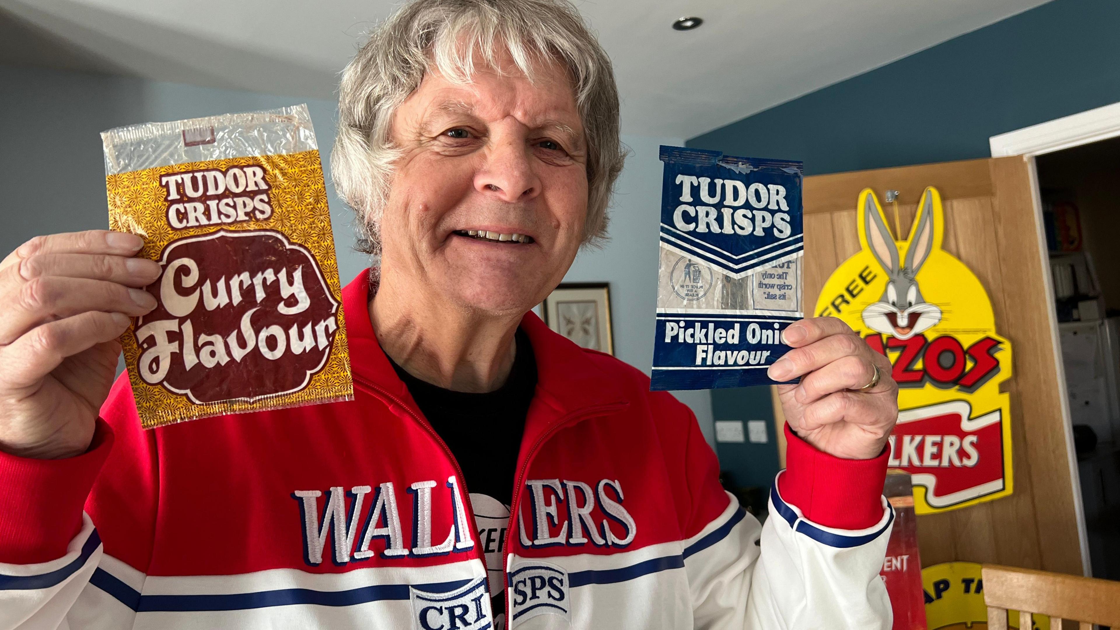 Frank Munford is holding an empty packet of vintage Tudor Crisps in each hand. On the left is a curry-flavoured packet and on the right is a pickled onion-flavour bag. He is holding them proudly, smiling at the camera. He is  wearing a vintage red and white Walkers Crisps tracksuit top.