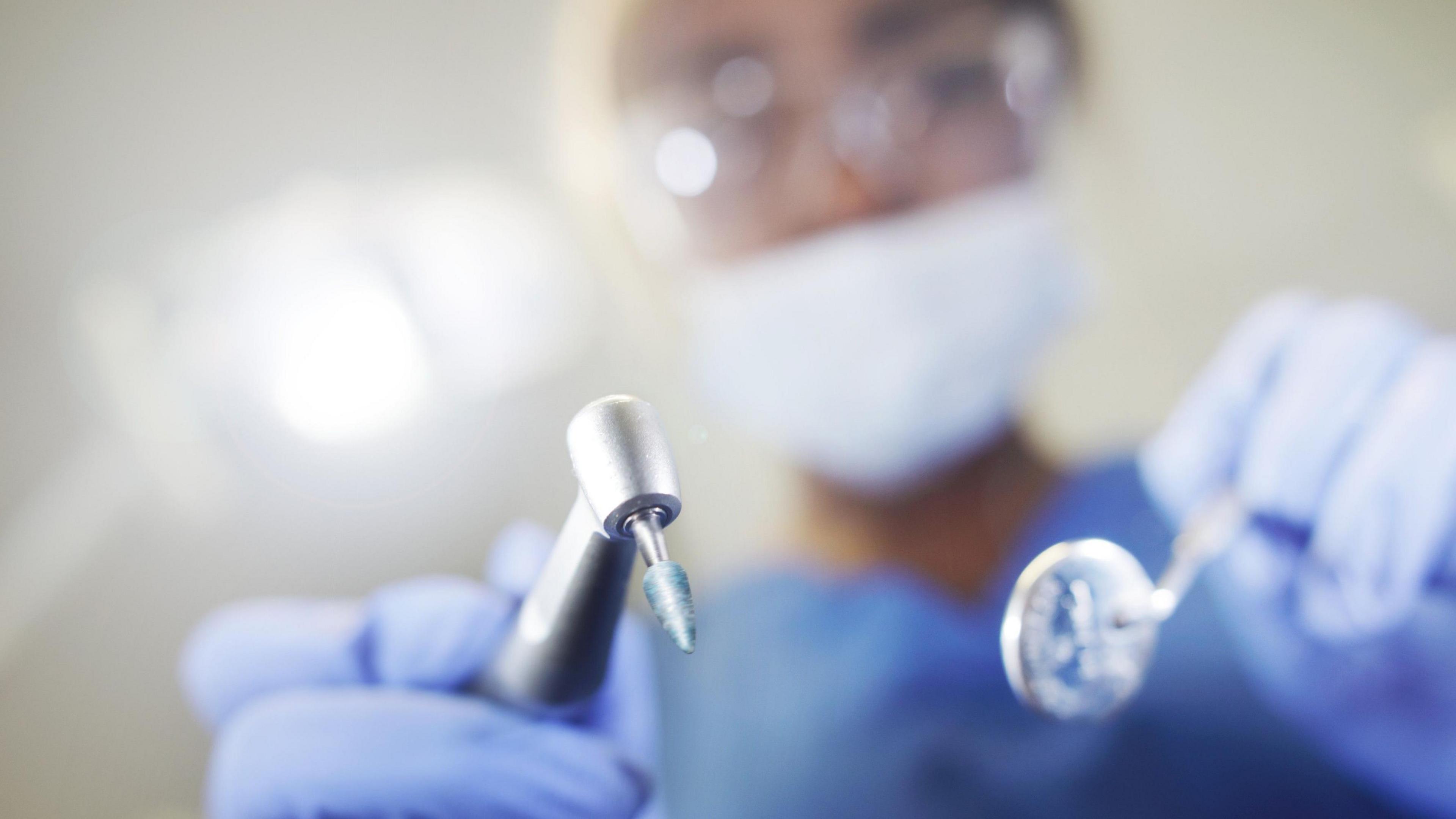 A dentist wearing PPE looks into the mouth of a patient, while holding a mirror and a drill.