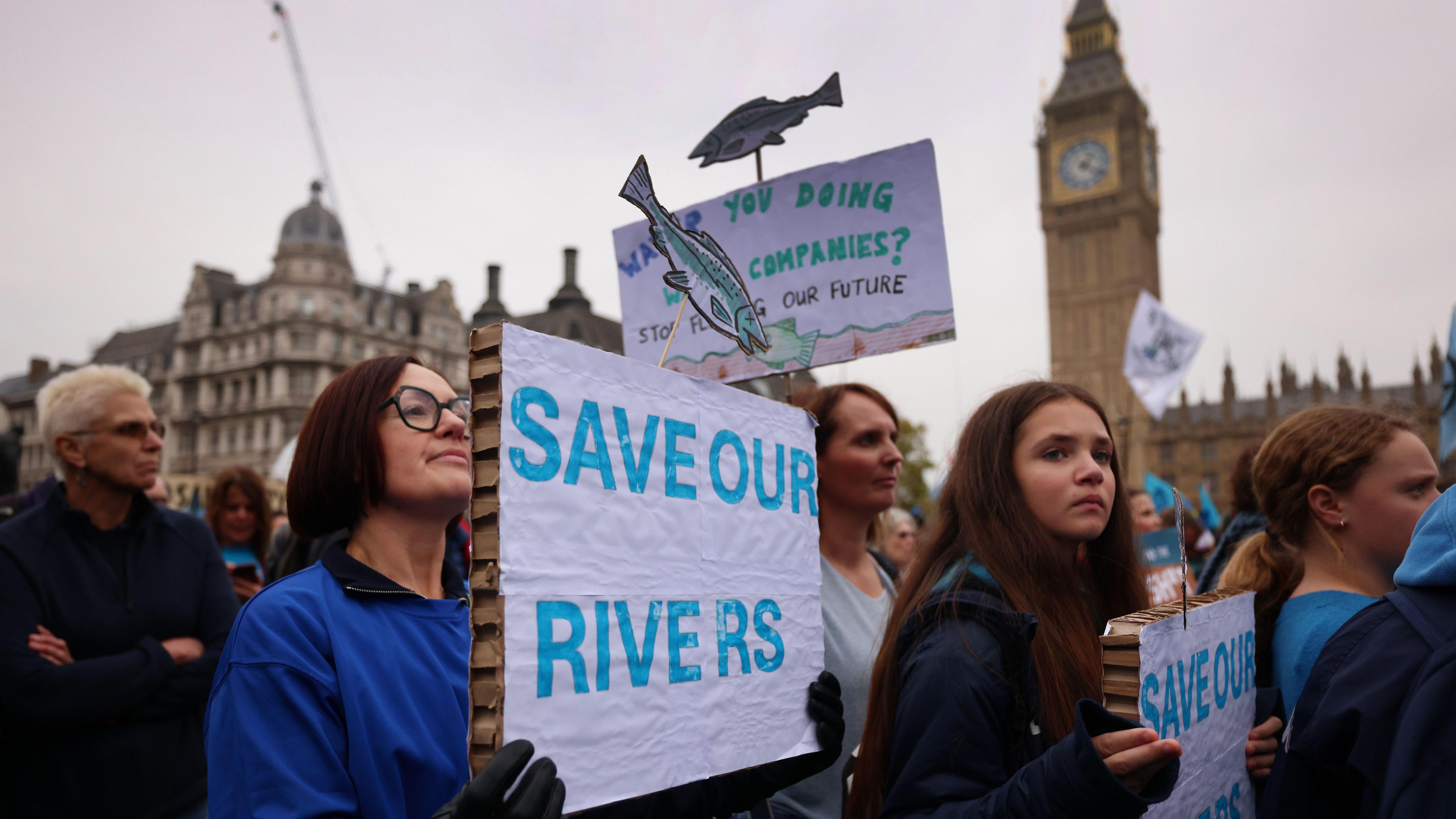 A crowd marching past the Elizabeth Tower near the Houses of Parliament. Two women are holding signs that say in blue writing Save Our Rivers.