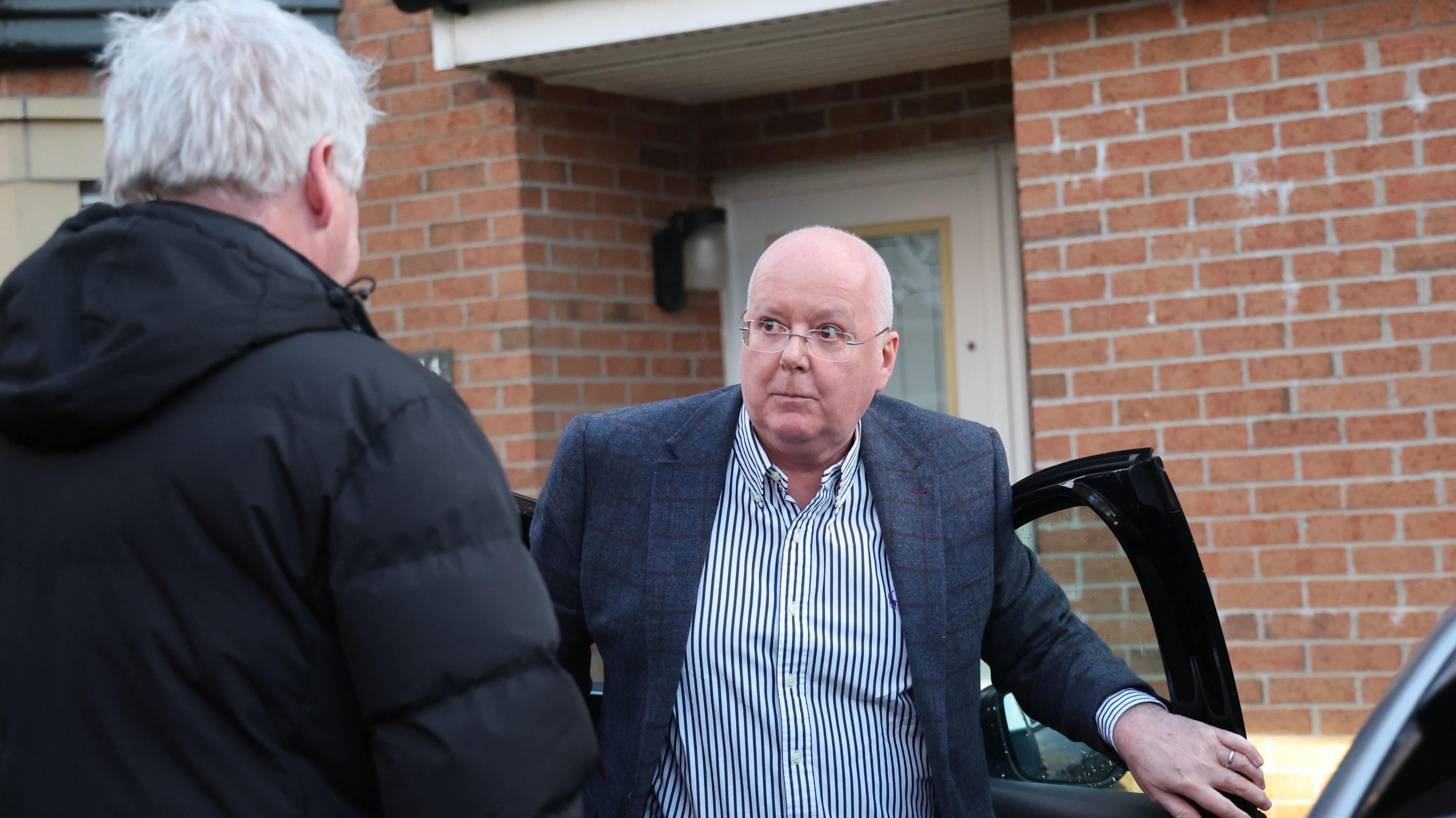 Peter Murrell in blue and white striped shirt and blue checked jacket exiting a car outside his modern brick home. A man is speaking to him, his back to the camera.