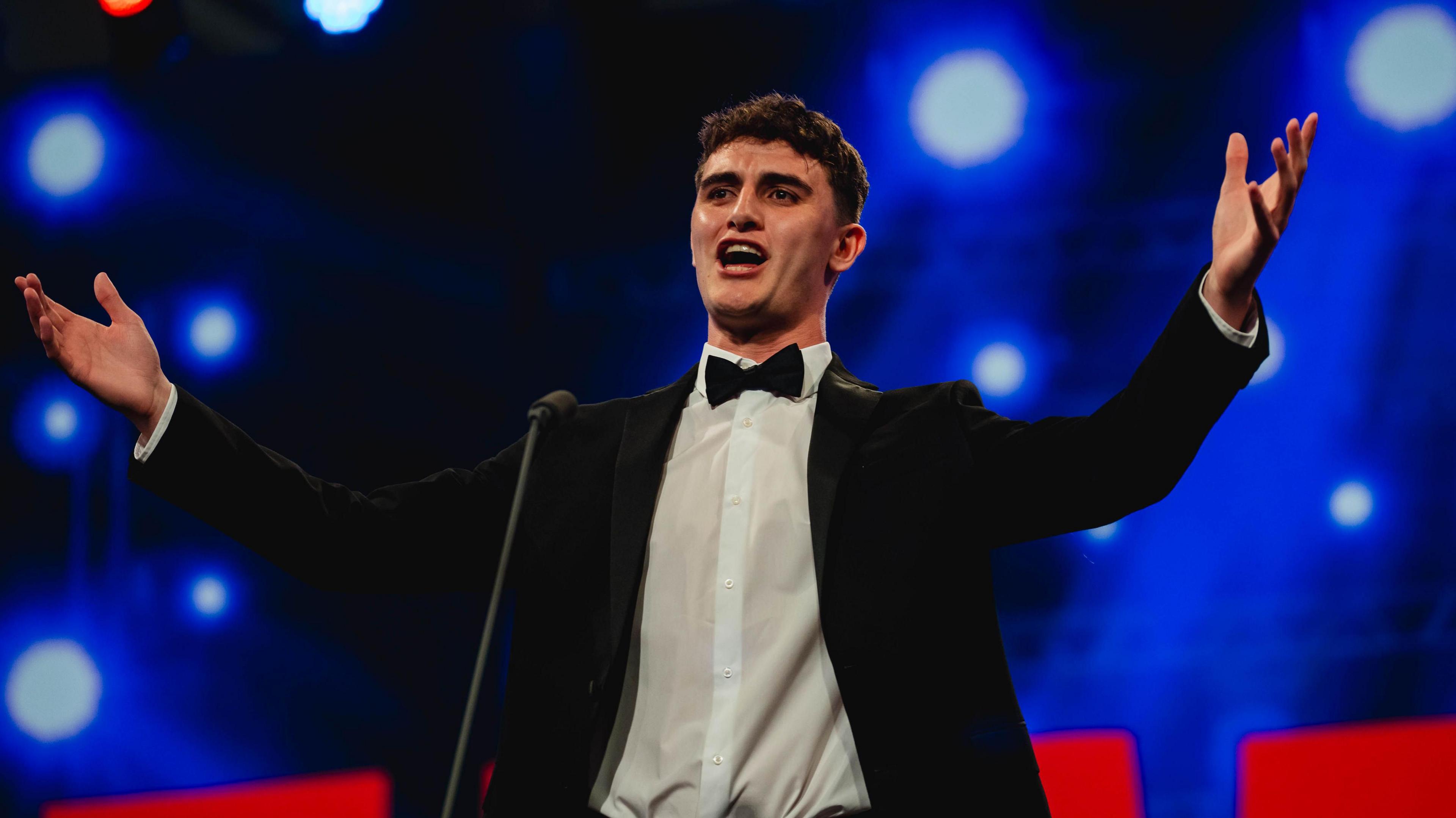 Caleb Nicholas stands on stage wearing a black suit and white shirt. His hands are outstretched as he sings. He has short dark hair.