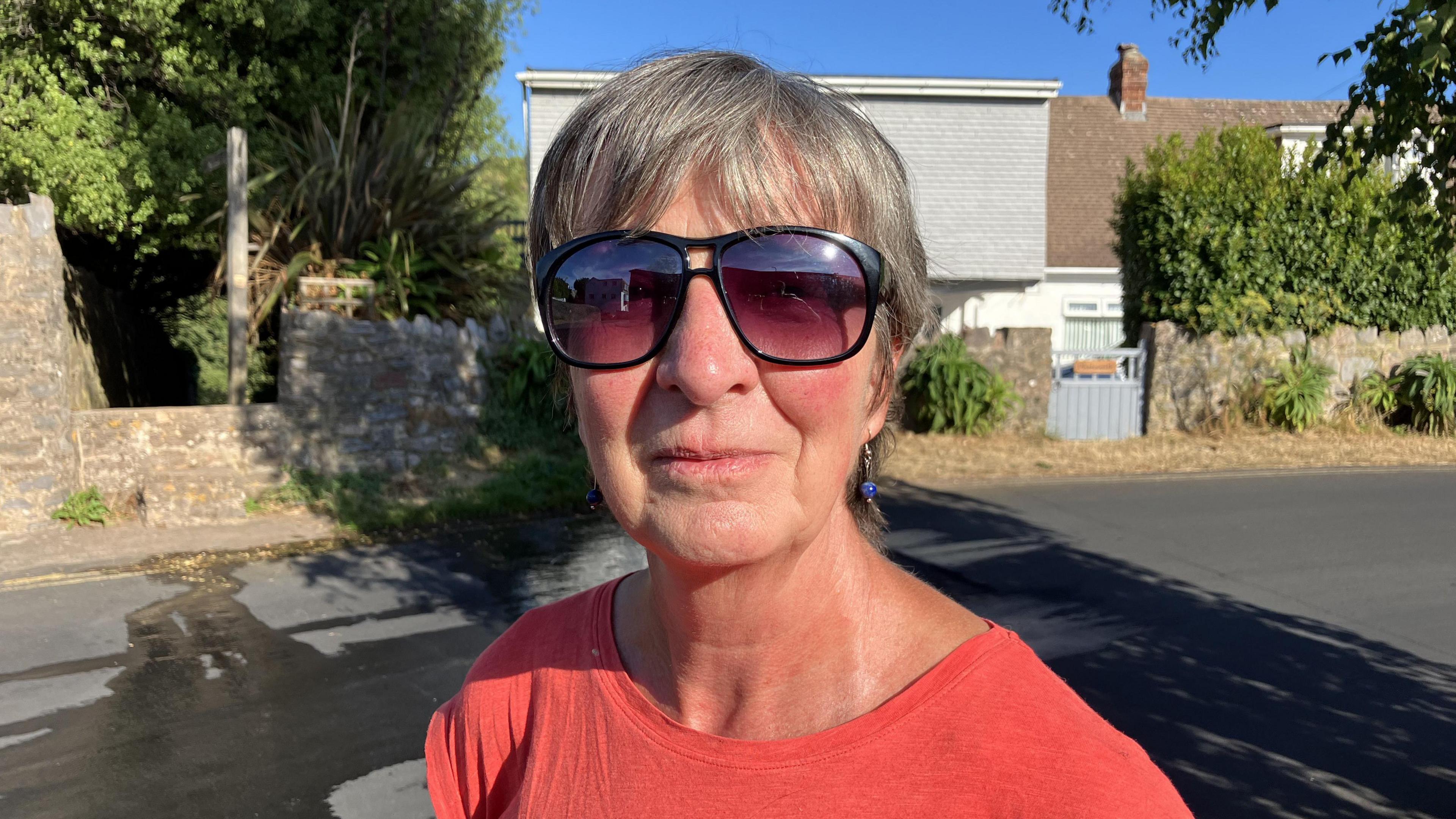Gillian Fisher - a woman in a red T-shirt and black sunglasses stands in Victoria Road in Brixham, in front of water that is running down the street from a leak on a verge.