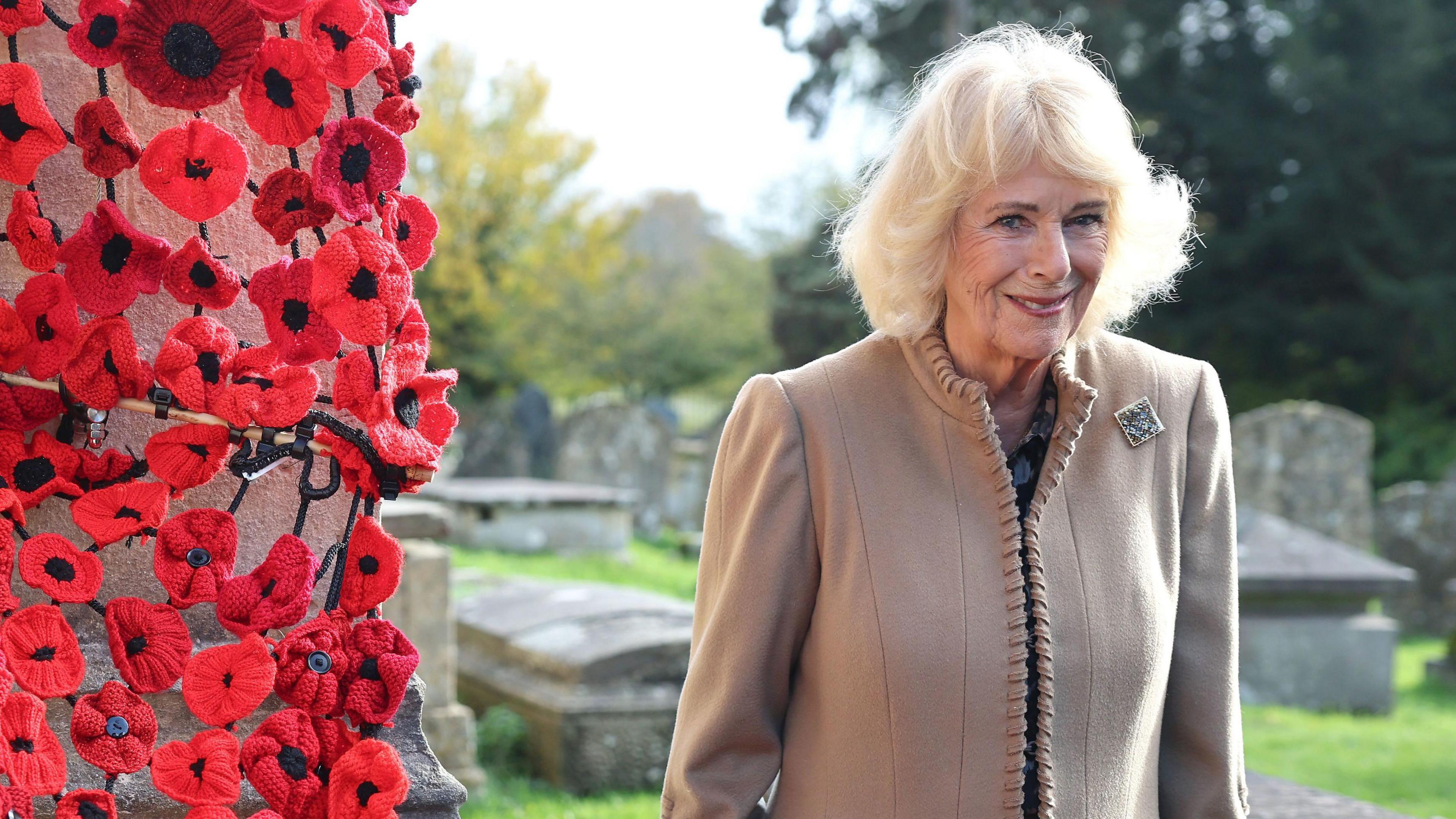 Queen Camilla in a brown jacket stood next to a large number of red knitted poppies