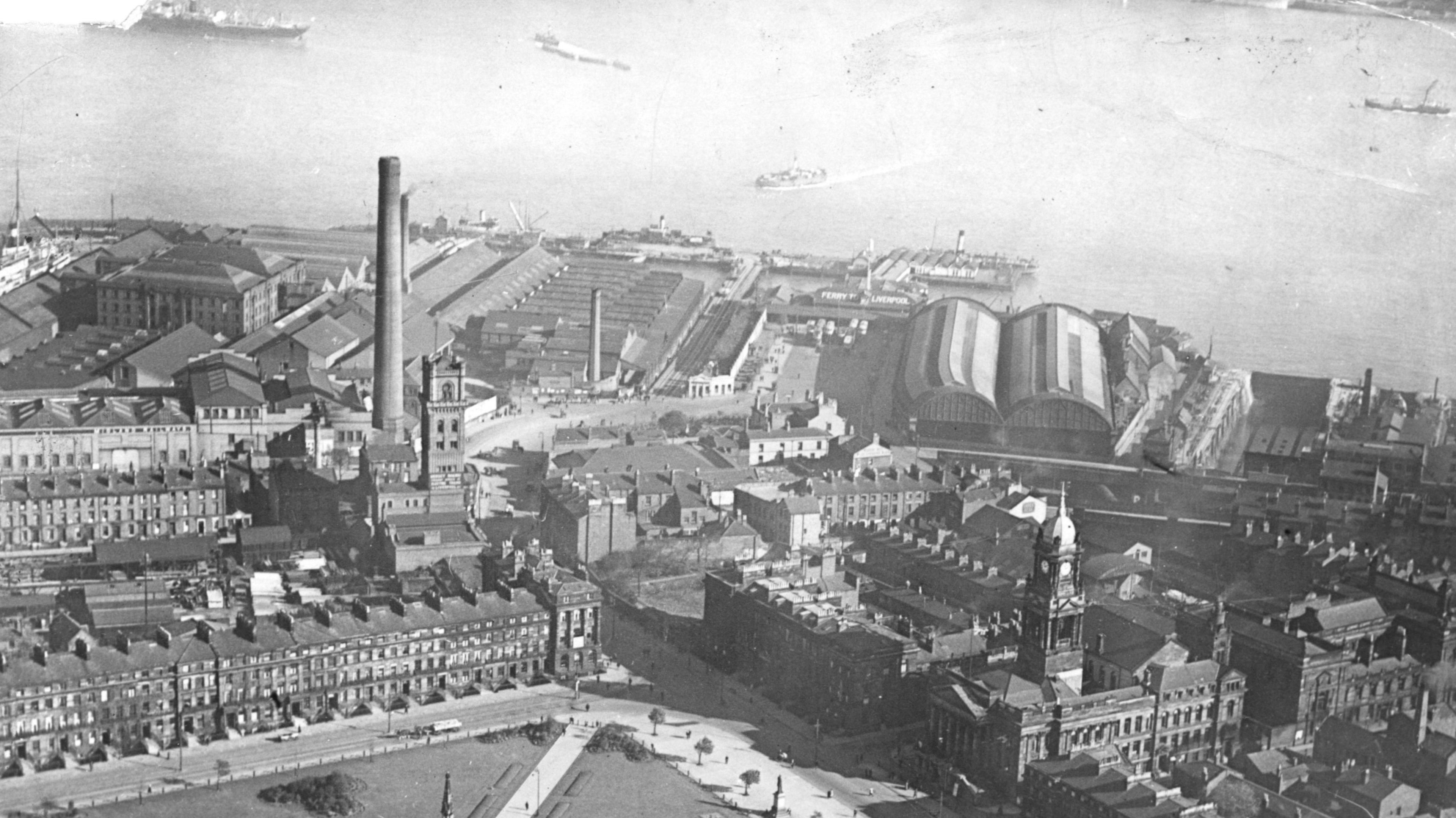A black and white aerial view of Hamilton Square surrounded by Georgian houses and a town hall. The roofs of the rail station platforms can be seen in the background by the River Mersey where a few vessels are sailing.