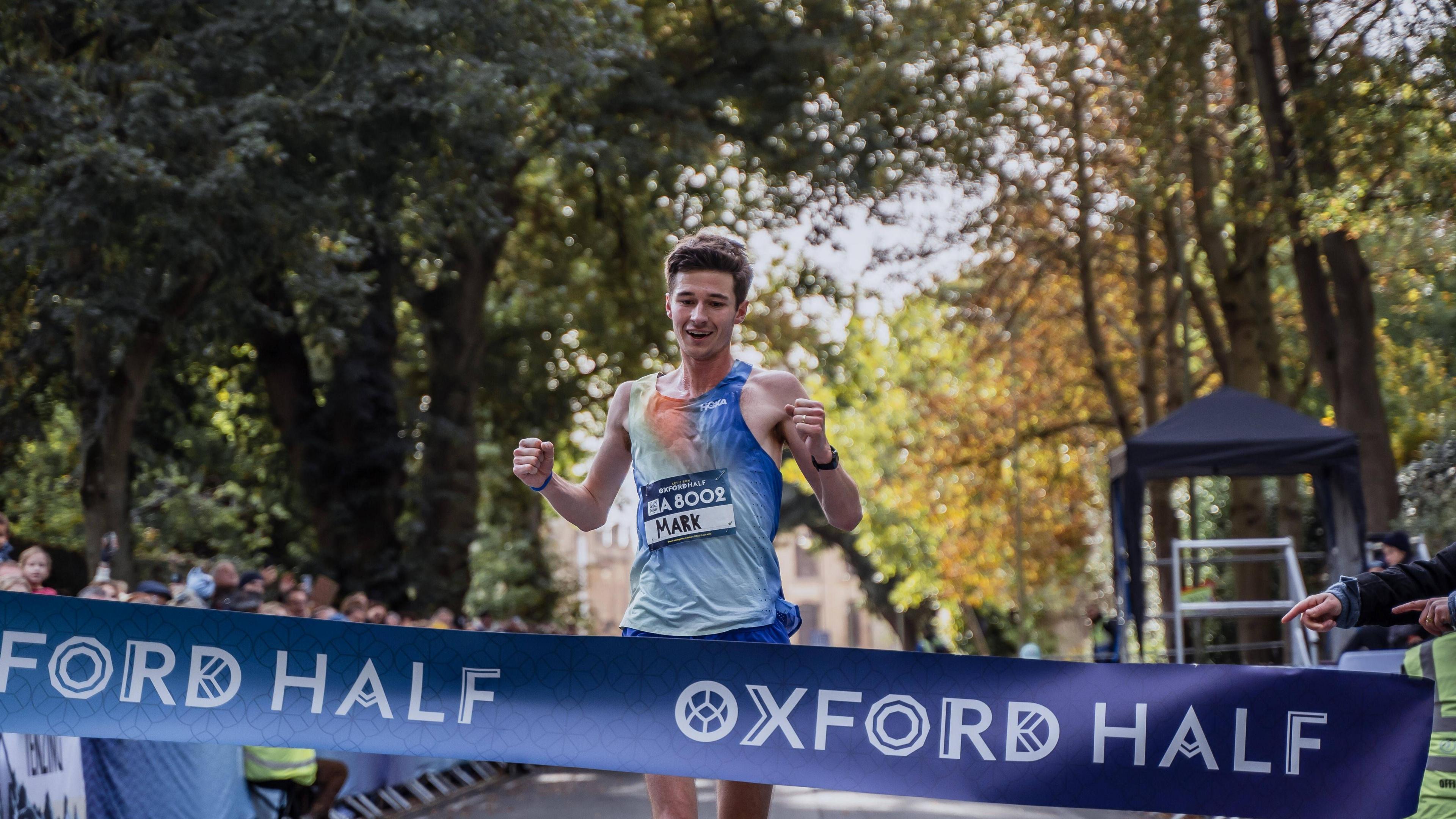 Mark Pearce crossing the finish line in first. He is wearing a blue running vest and there is a banner saying 'oxford half' in front of him.