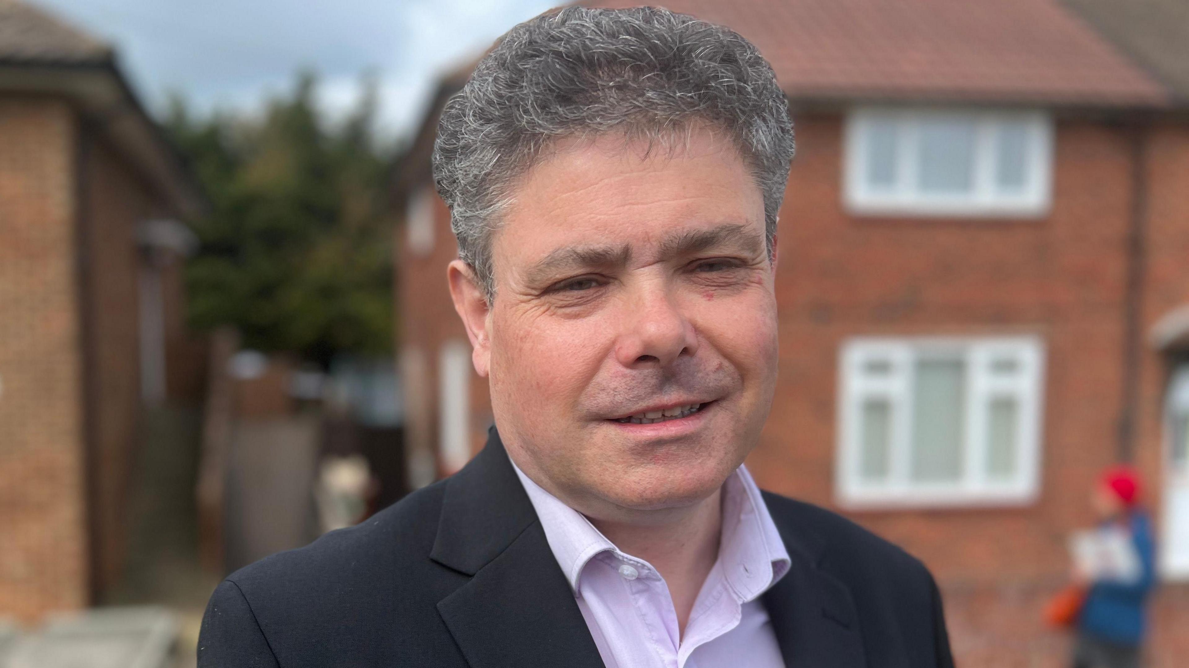 John Kent looks directly at the camera dressed in a suit and shirt, he is standing in front of a residential house which is blurred. 